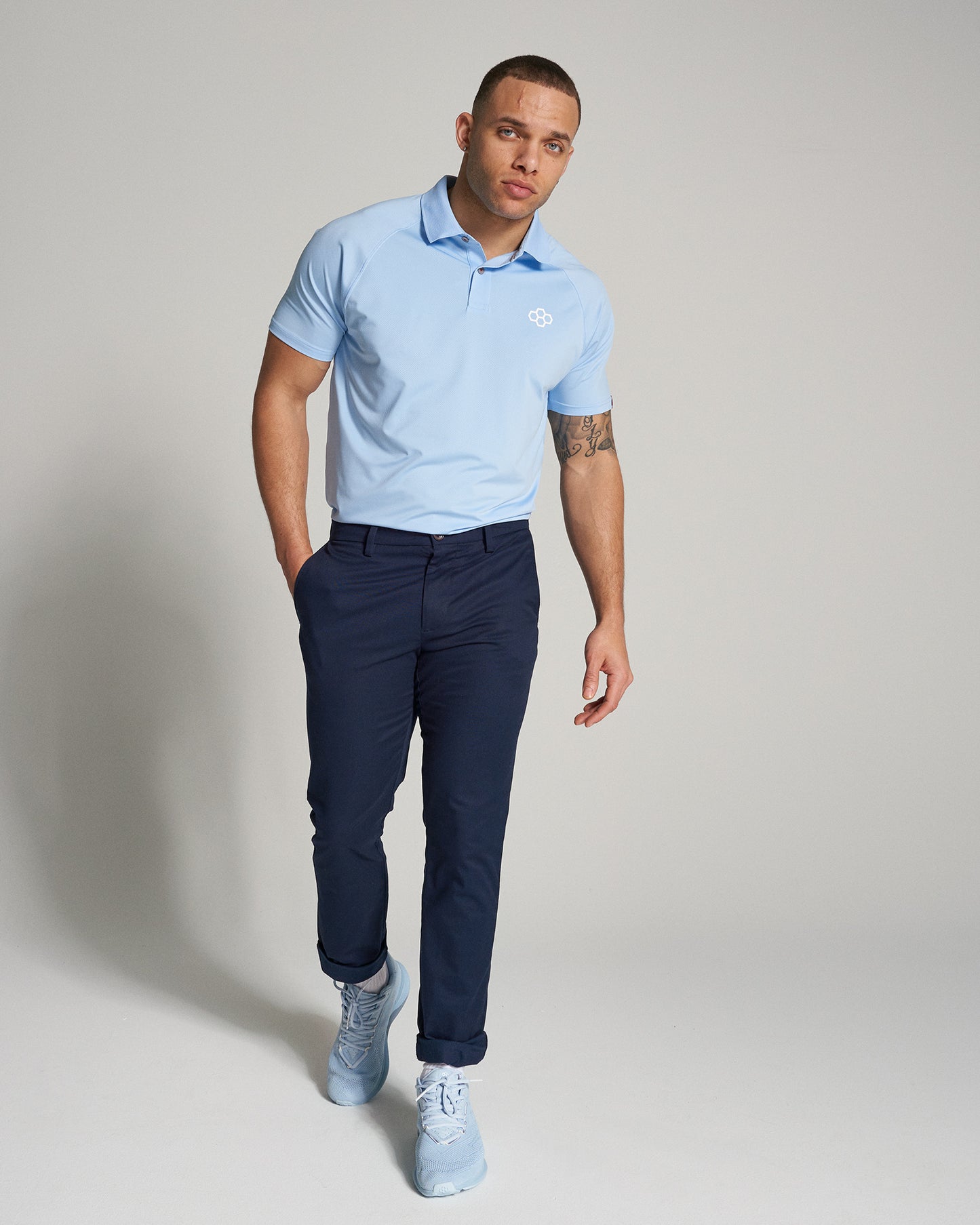 A stylish man is confidently walking in a light blue polo shirt dark pants and light blue sneakers posing against a neutral background