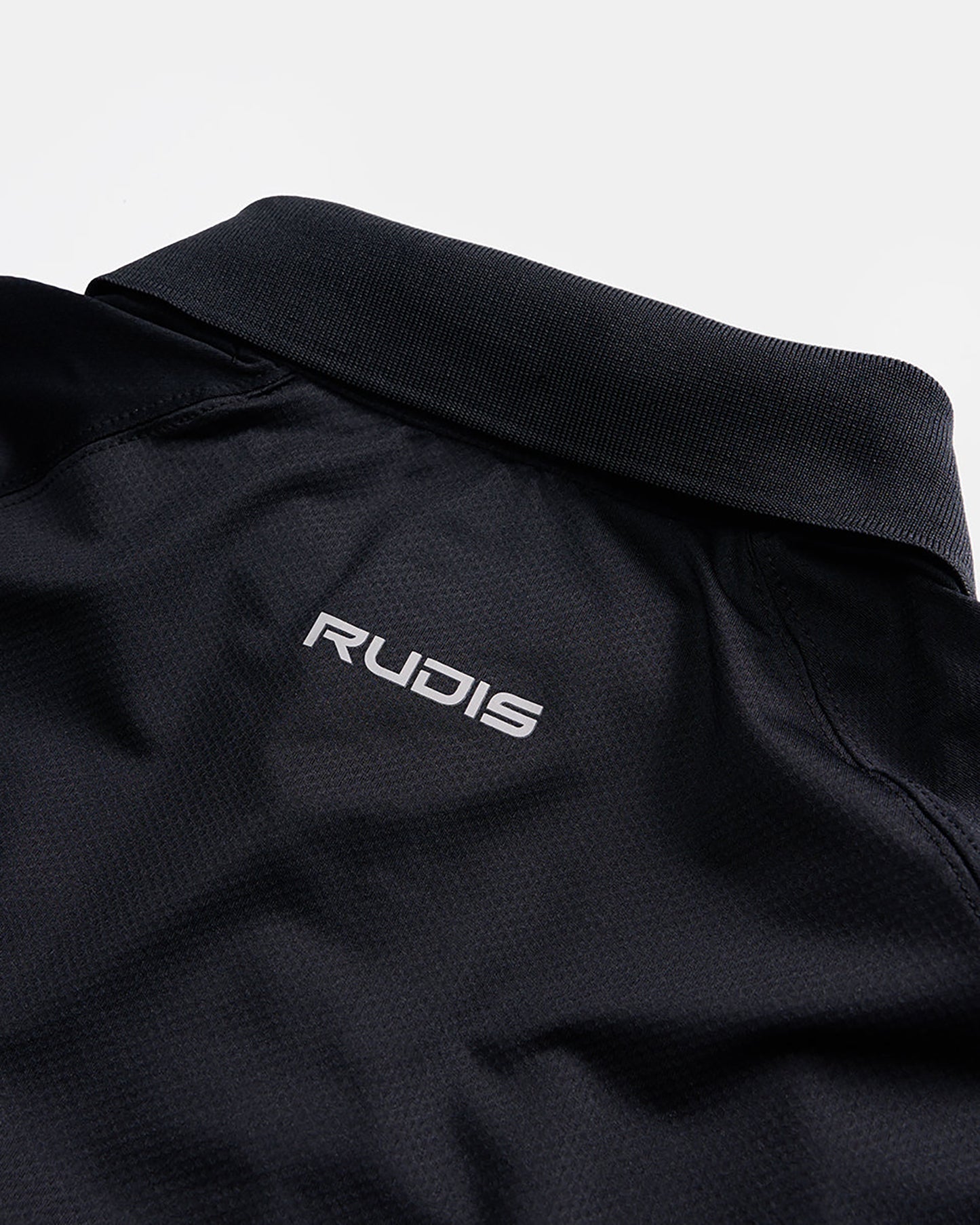 A close-up view of a black athletic polo shirt featuring a slim structured collar and a distinctive logo that reads RUDIS