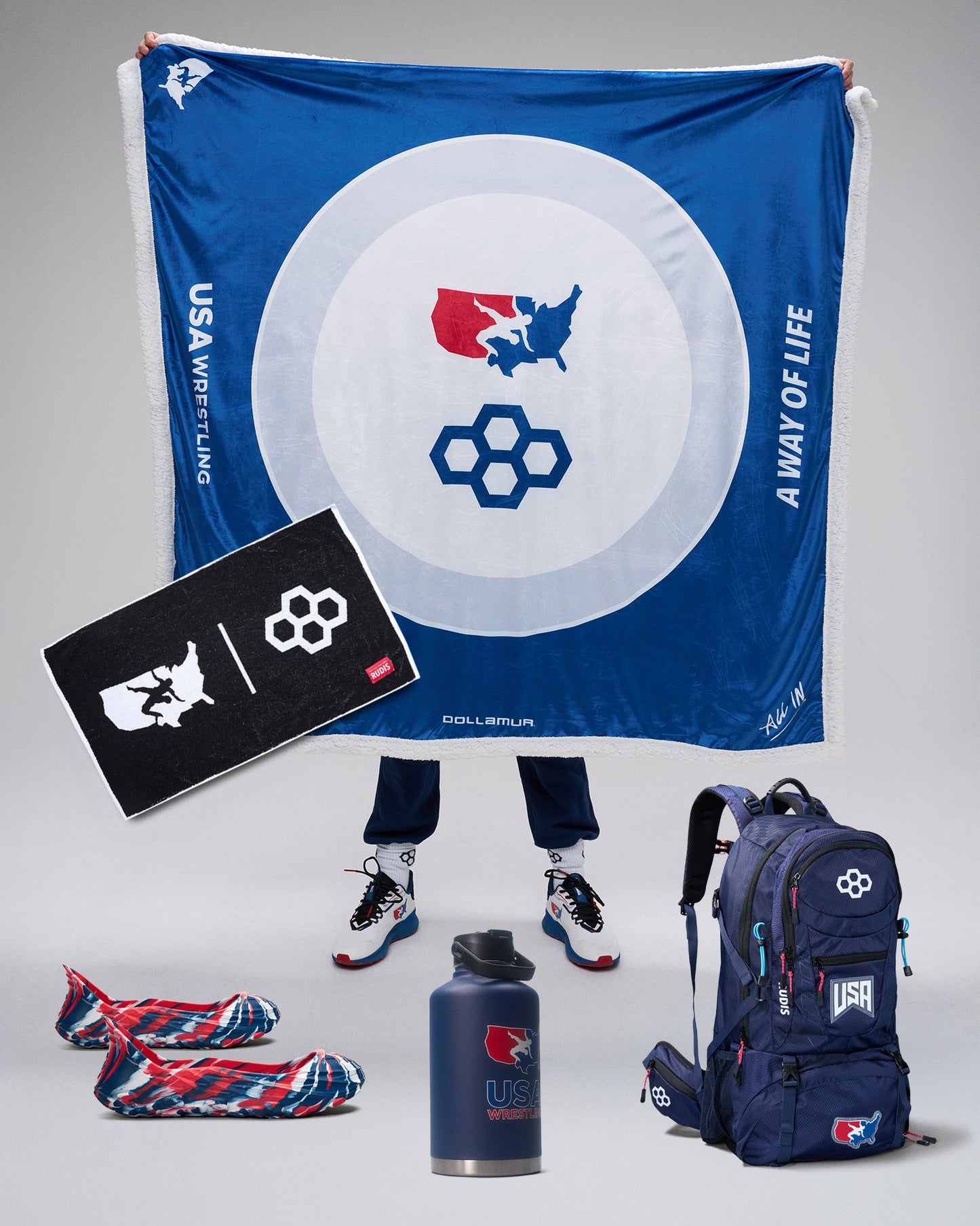 USA Wrestling branded merchandise including a blanket, backpack, towel, and water bottle on a gray background.