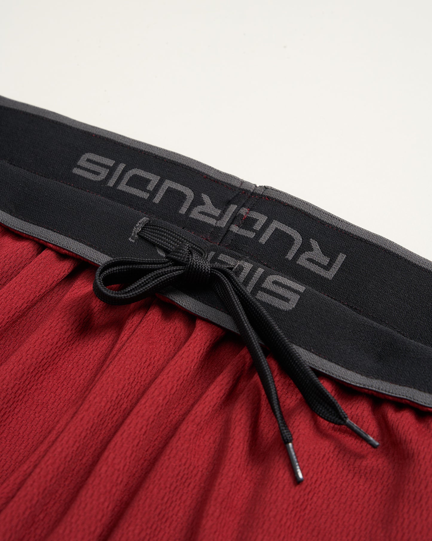 This image displays a close-up view of red athletic shorts featuring a black elastic waistband with the brand name printed on it and contrasting drawstrings for a secure fit