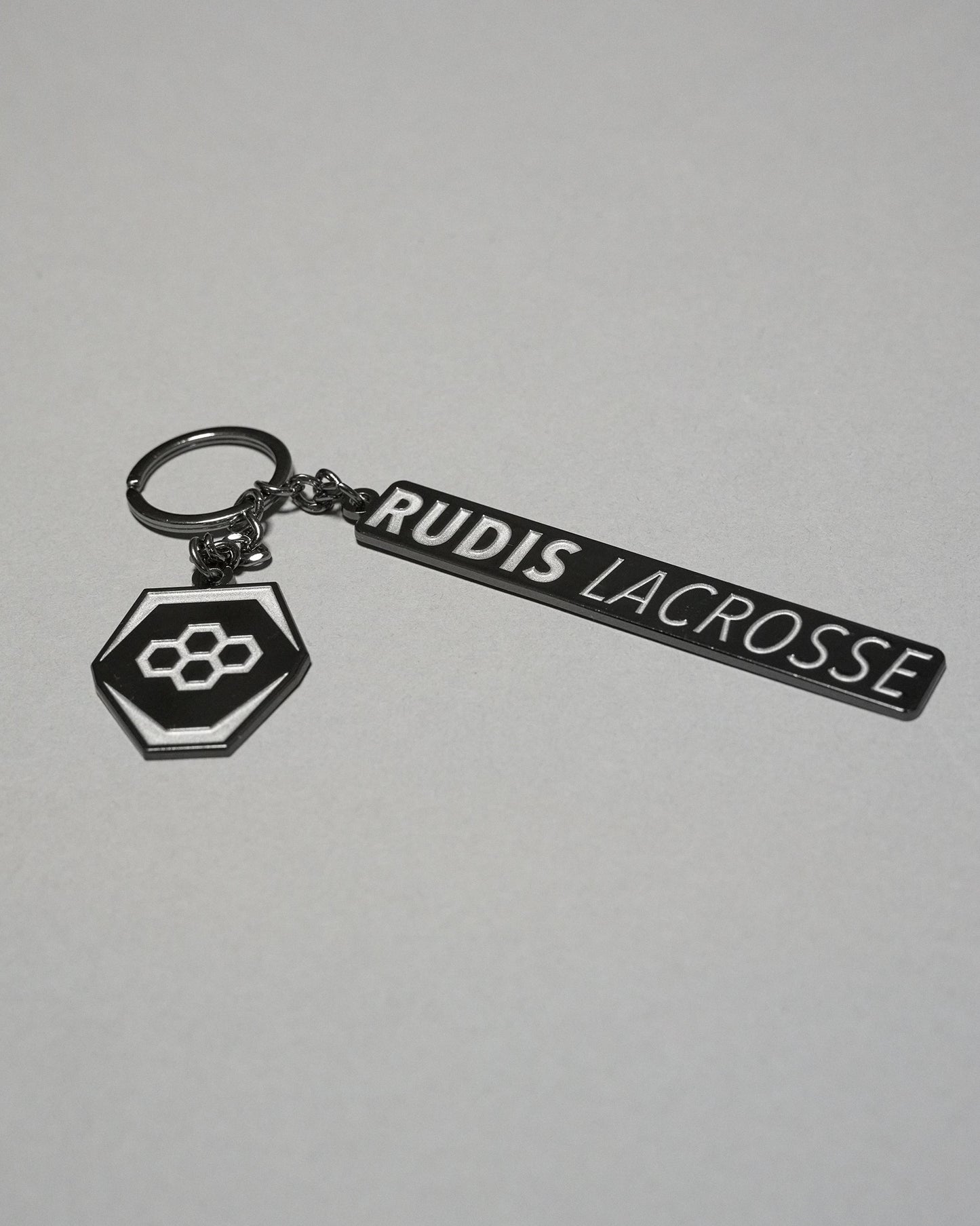 RUDIS Lacrosse metal keychain in black and silver featuring the signature RUDIS hex logo, engraved RUDIS Lacrosse branding, durable metal construction, and split key ring.