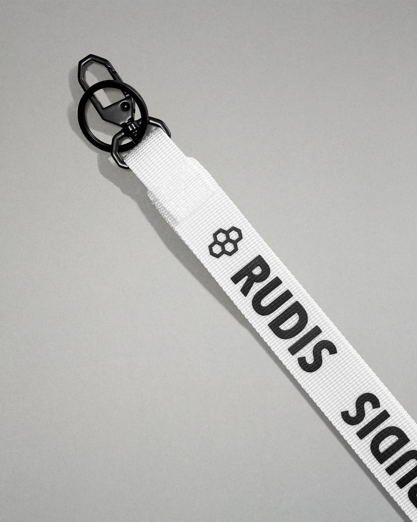 Close-up of the white RUDIS Wordmark lanyard featuring a durable metal key clip, key ring, woven polyester strap, and bold white RUDIS logo with honeycomb icon.
