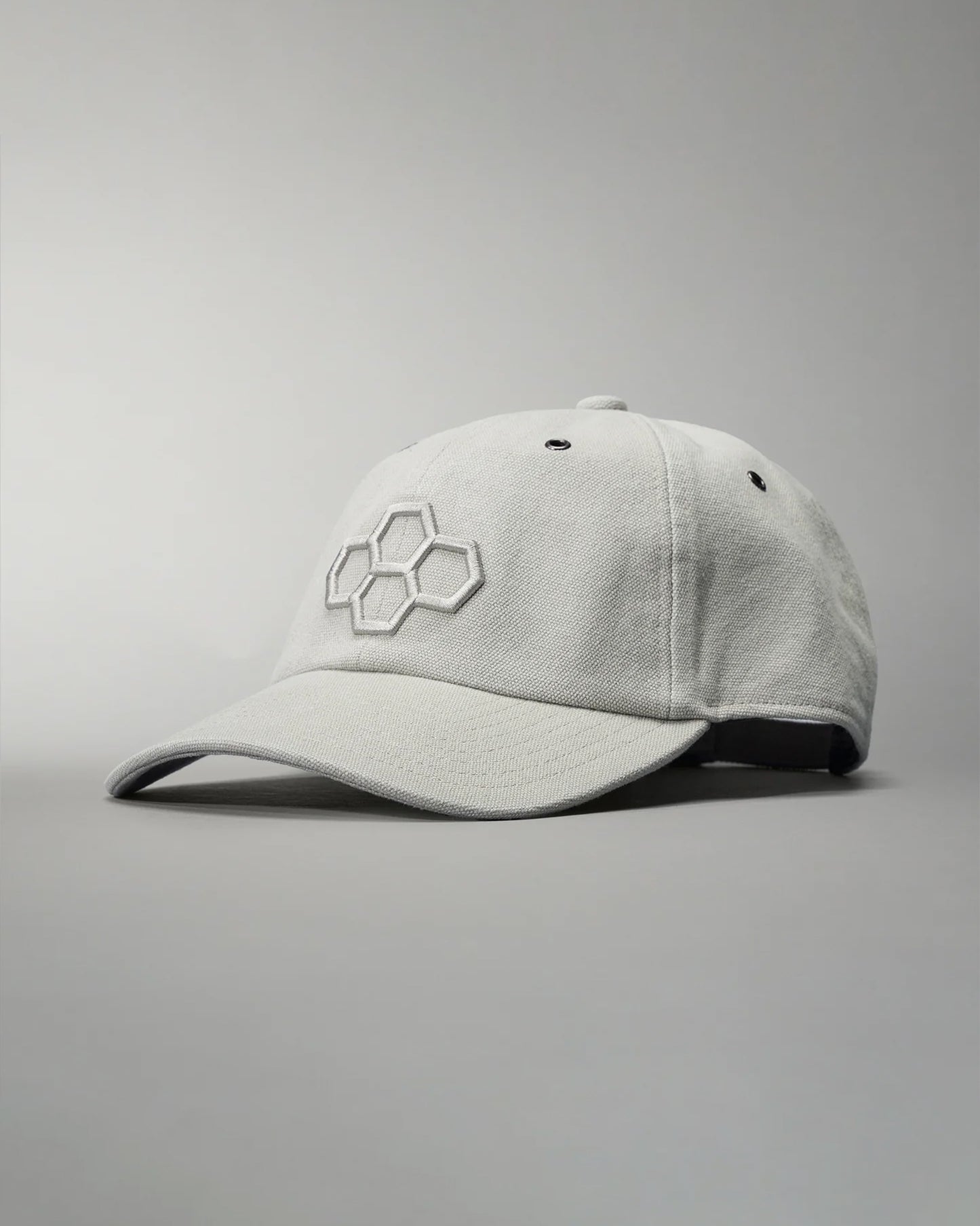 A light gray baseball cap with an embroidered hexagonal logo is displayed at an angle against a muted gray background, highlighting its structured design and comfortable fabric.