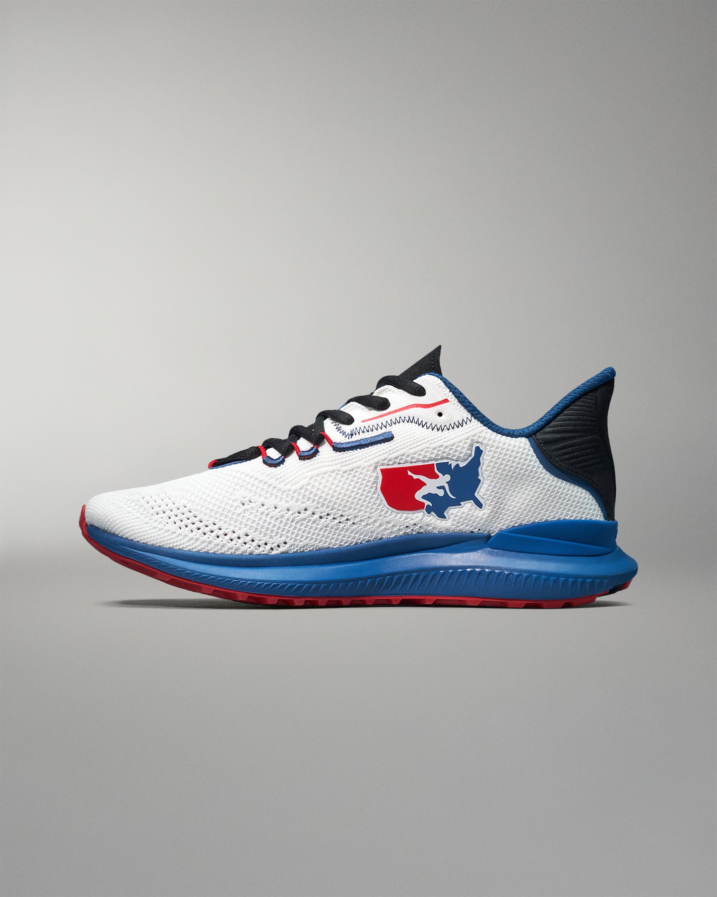 Side view of a white RUDIS training shoe with blue and red detailing, featuring the USA Wrestling logo on the heel, black laces, and a blue midsole with red tread, displayed against a neutral gray background.