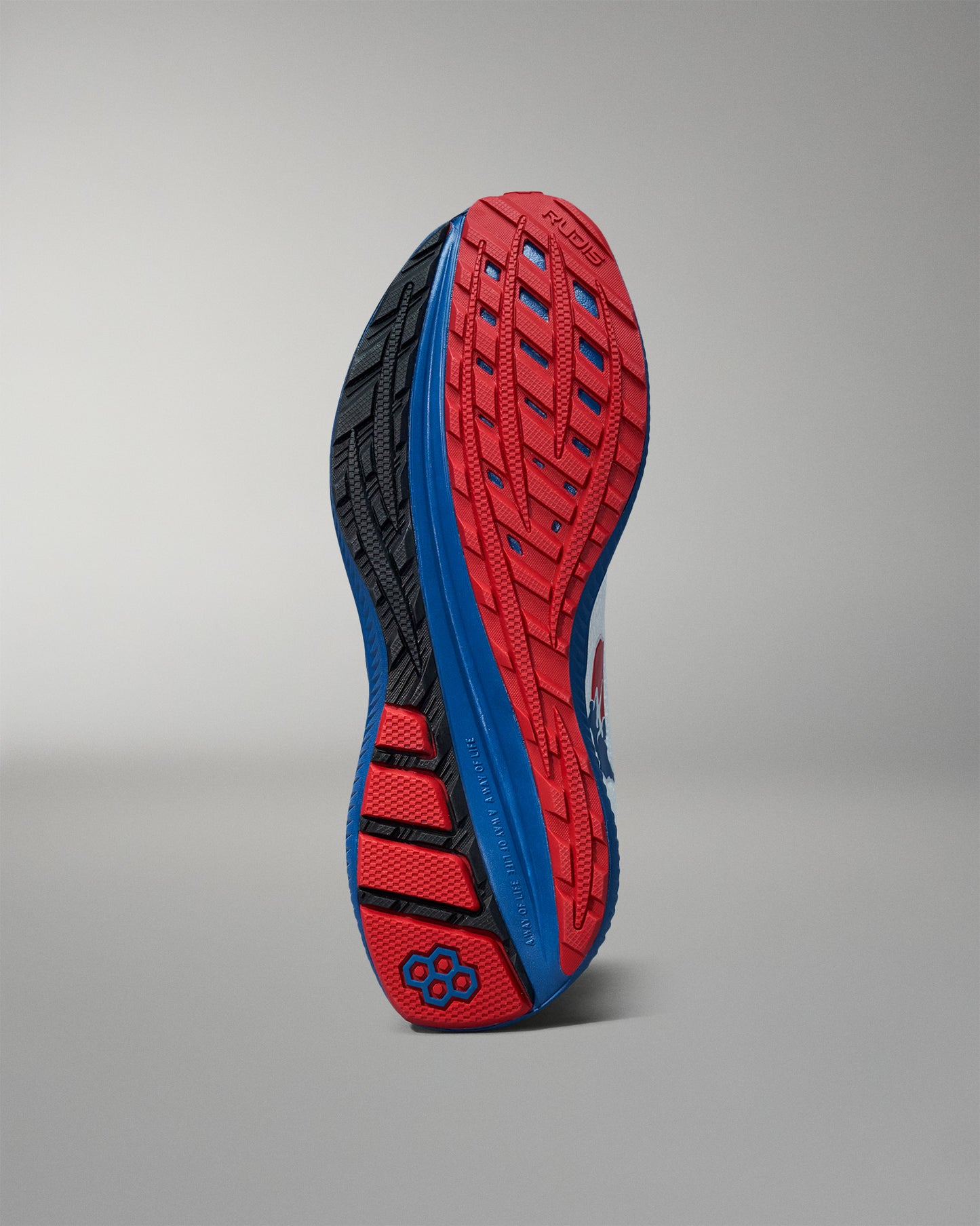 Bottom view of a RUDIS training shoe showcasing a red, blue, and black high-traction outsole with a hexagon logo near the heel. Displayed against a neutral gray background.