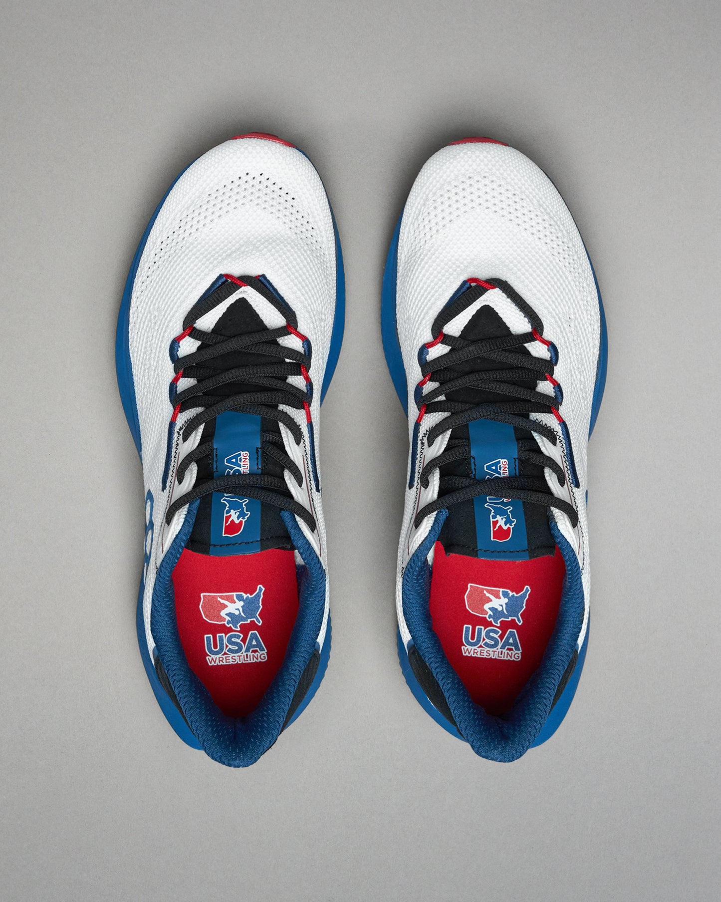 Top view of a pair of white RUDIS training shoes with black laces, blue and red accents, and USA Wrestling logos visible on the red insoles and blue tongues. Displayed on a light gray background.