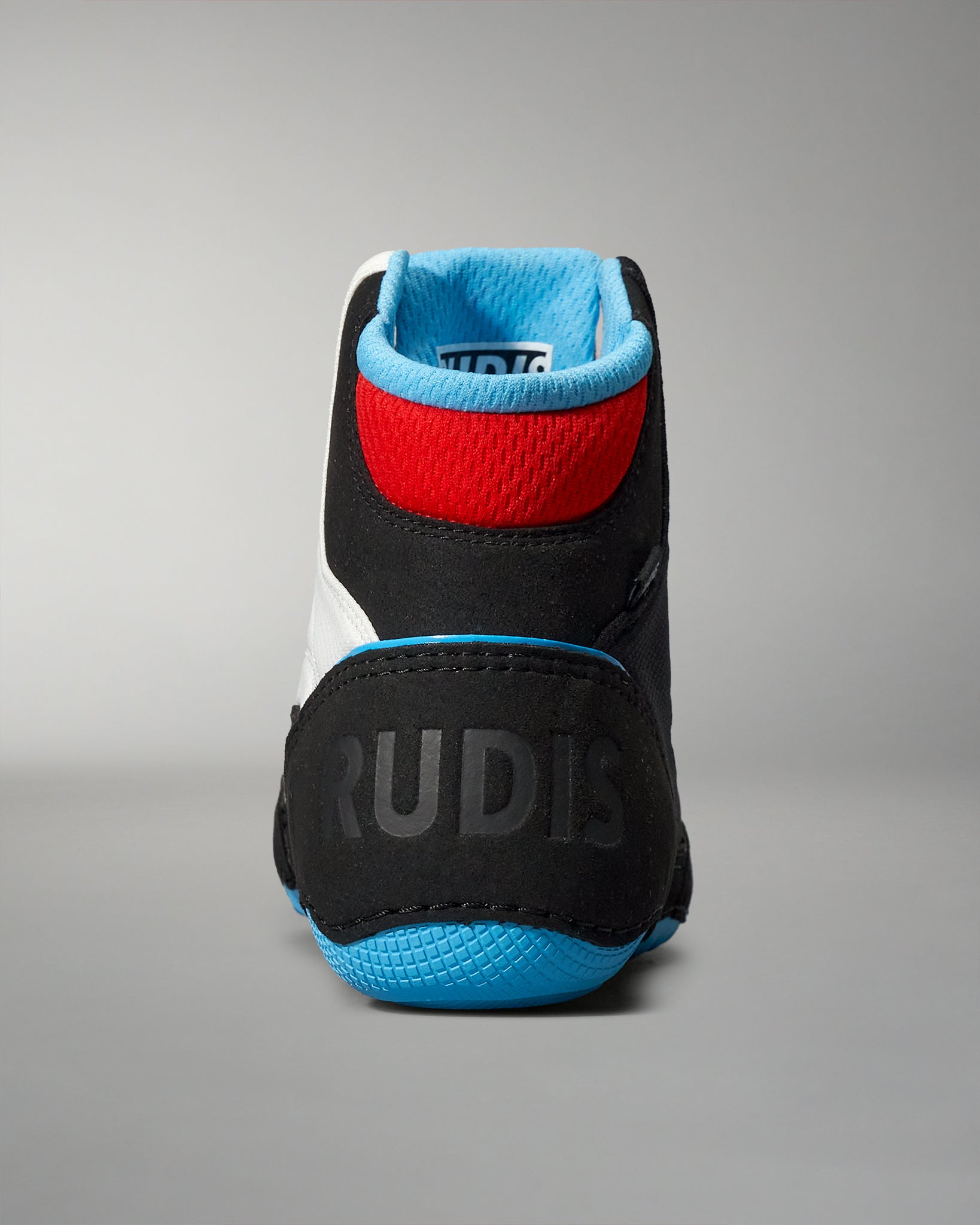 Back view of a wrestling shoe in black and white, accented with red and blue, featuring 'RUDIS' on the textured heel against a neutral gray background.
