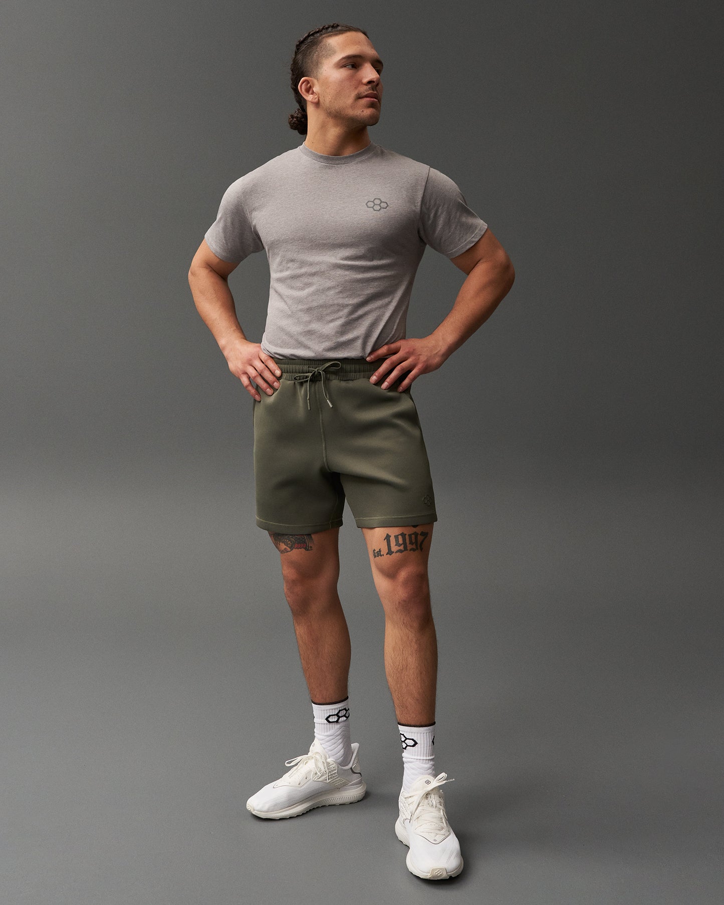 A fit man poses confidently in a gray t-shirt and olive green shorts complemented by white athletic shoes and patterned socks against a muted background