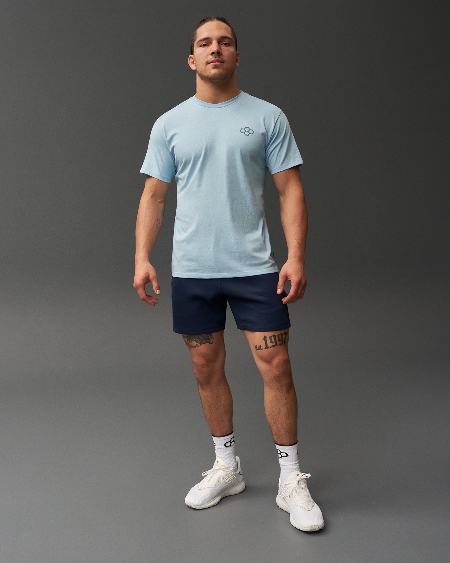 A fit man poses confidently wearing a light blue t-shirt dark shorts and white athletic shoes showcasing a stylish and active look