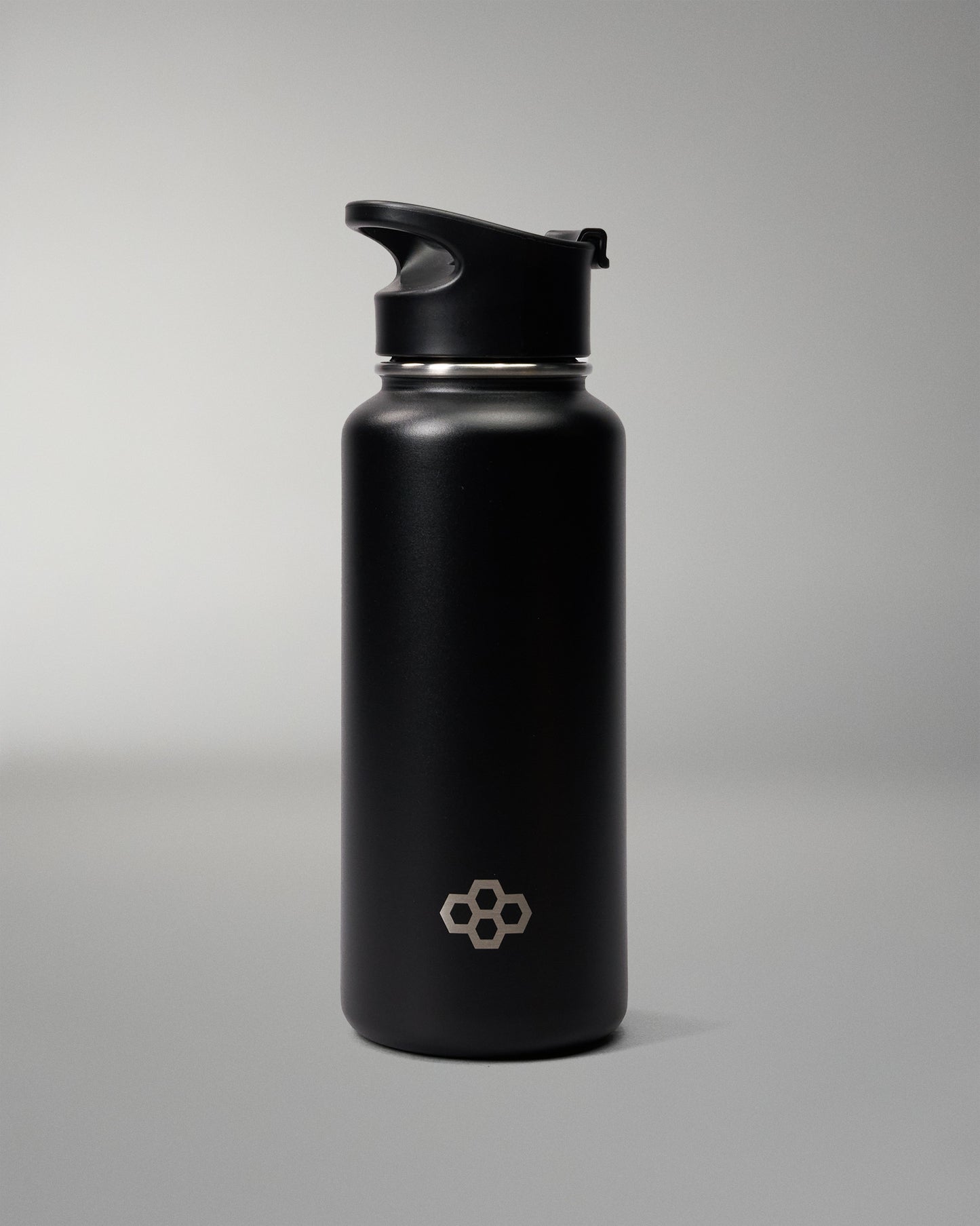A matte black insulated water bottle with a matching black flip-top lid, featuring a small silver geometric logo near the bottom, set against a neutral gray studio background.