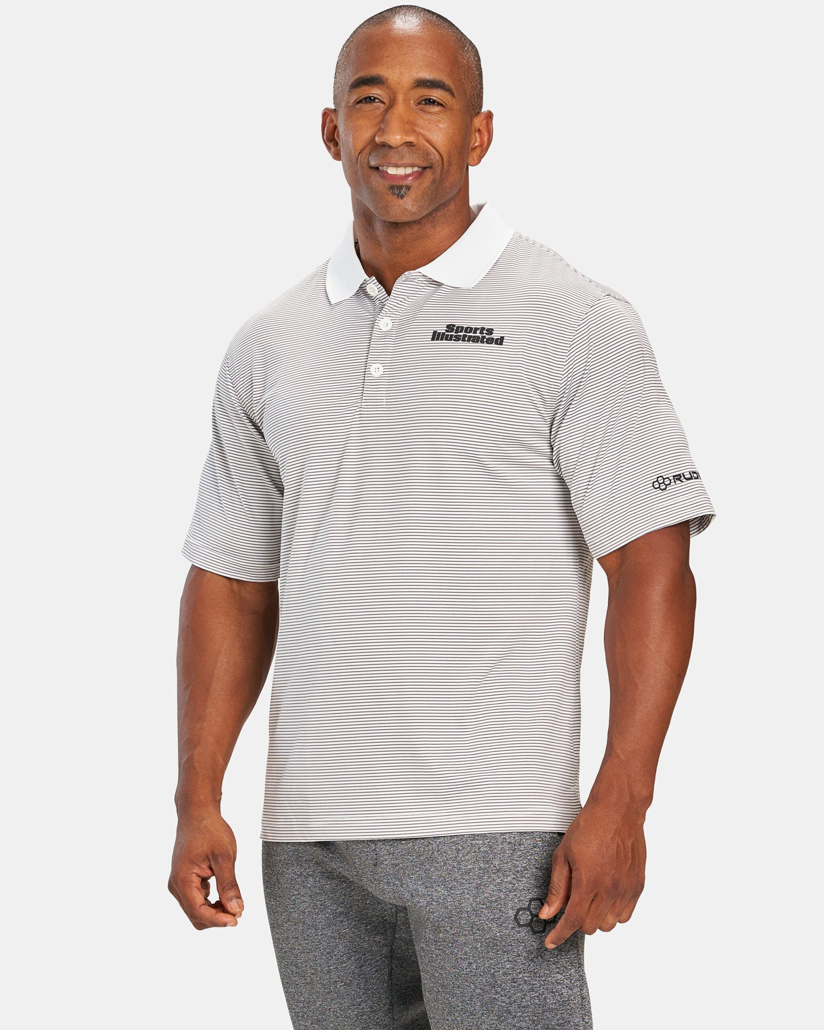A man smiles while wearing a short-sleeved striped polo shirt with the Sports Illustrated logo paired with gray athletic pants
