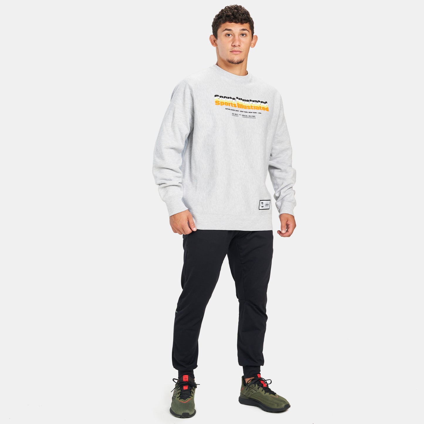 A young man stands confidently in a casual outfit featuring a light gray sweatshirt with Sports Illustrated printed on it black tapered pants and olive green sneakers set against a minimalist background