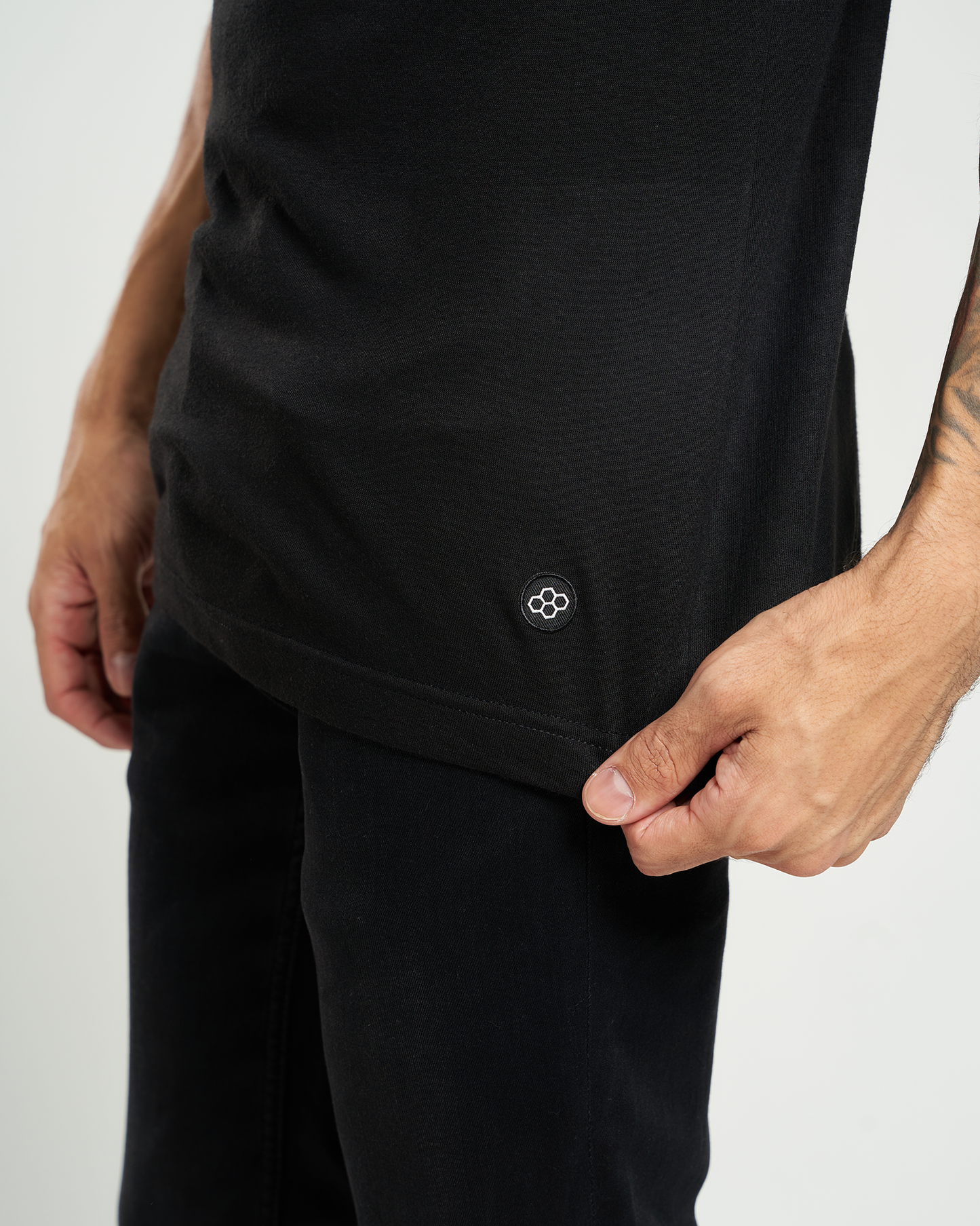 A close-up view of a man holding the hem of a black shirt revealing a small embroidered logo at the bottom