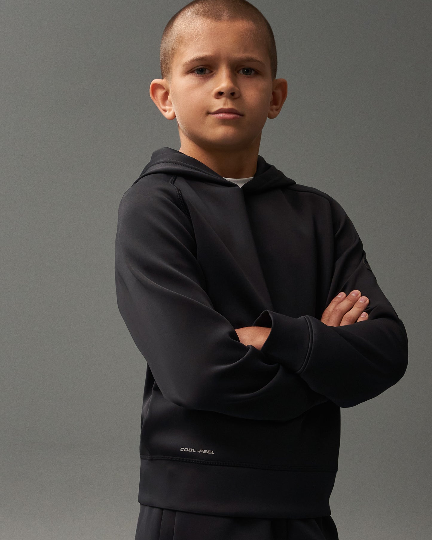 A young boy confidently poses with crossed arms in a black hooded sweatshirt, featuring a 'COOL-FEEL' logo, against a neutral background.