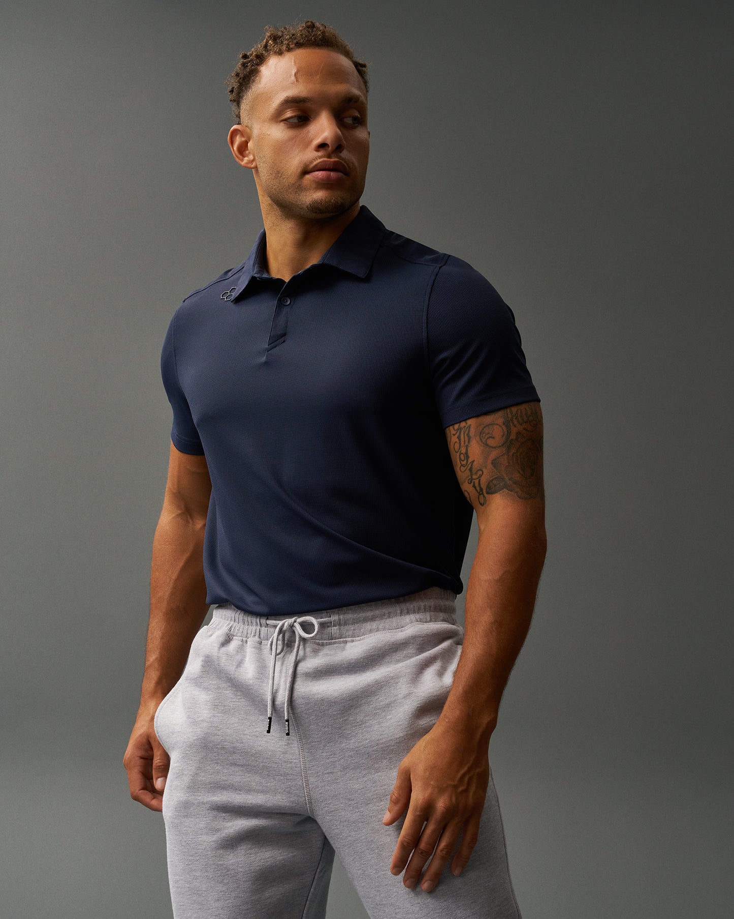 A man stands confidently against a gray background wearing a fitted navy RUDIS polo shirt and light gray drawstring sweatpants. The polo features a subtle hexagonal RUDIS logo near the collar. He has a muscular build, visible tattoos, and looks slightly to his left.