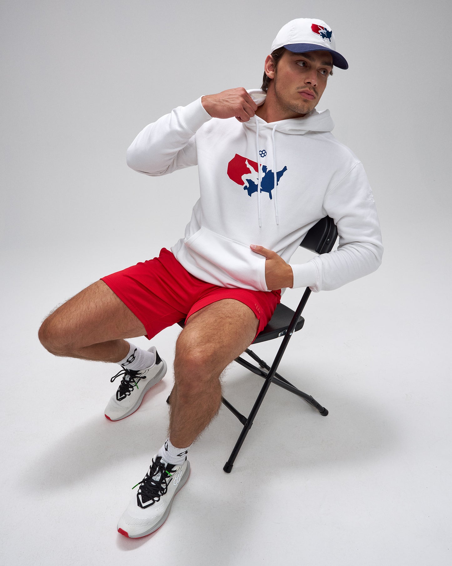 Person wearing a white hoodie with a logo, red shorts, and white sneakers on a gray background