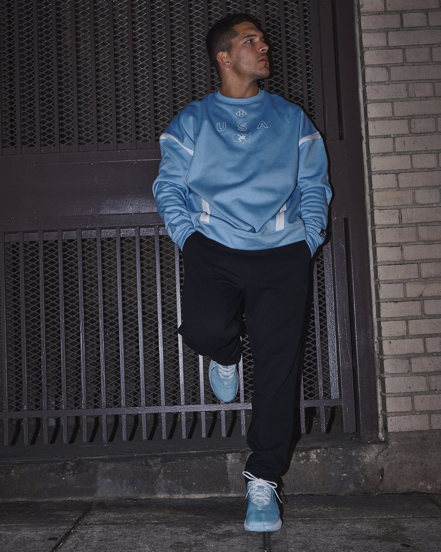 Person wearing a blue sweatshirt and black pants leaning against a metal gate.