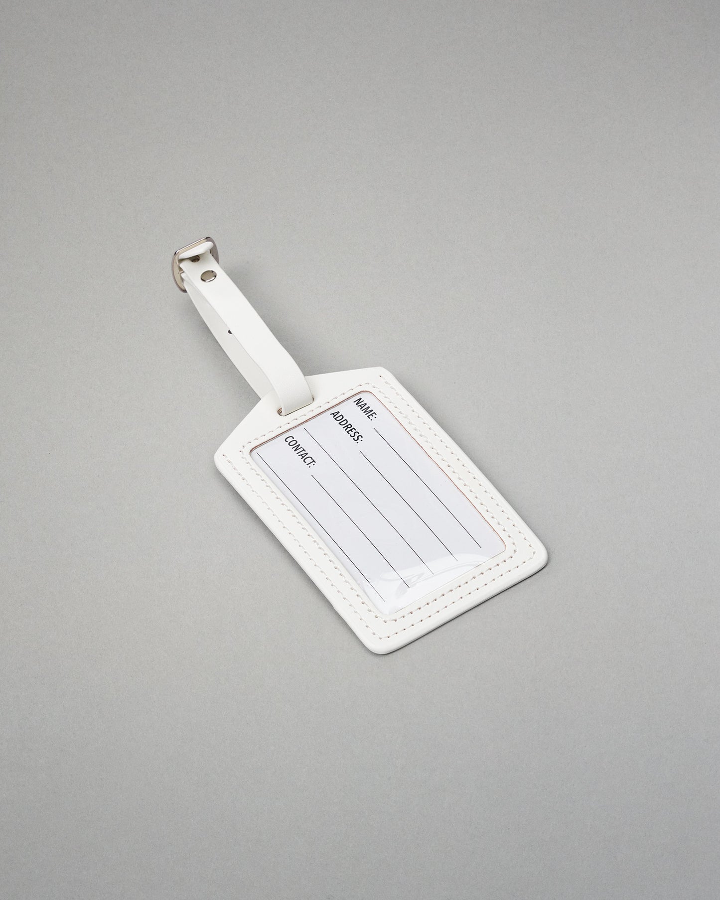 A white leather luggage tag with a silver metal buckle, featuring blank lines for name, address, and contact information, set against a solid gray background.