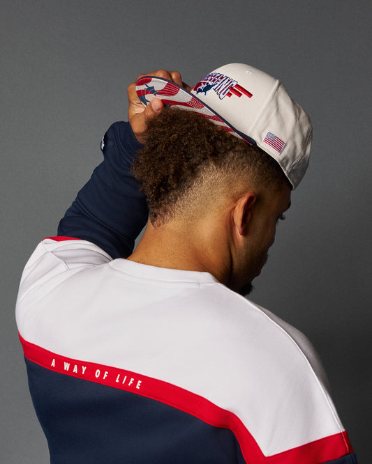 Person wearing a white cap with red and blue design, and a white and navy jacket with red text on a gray background.
