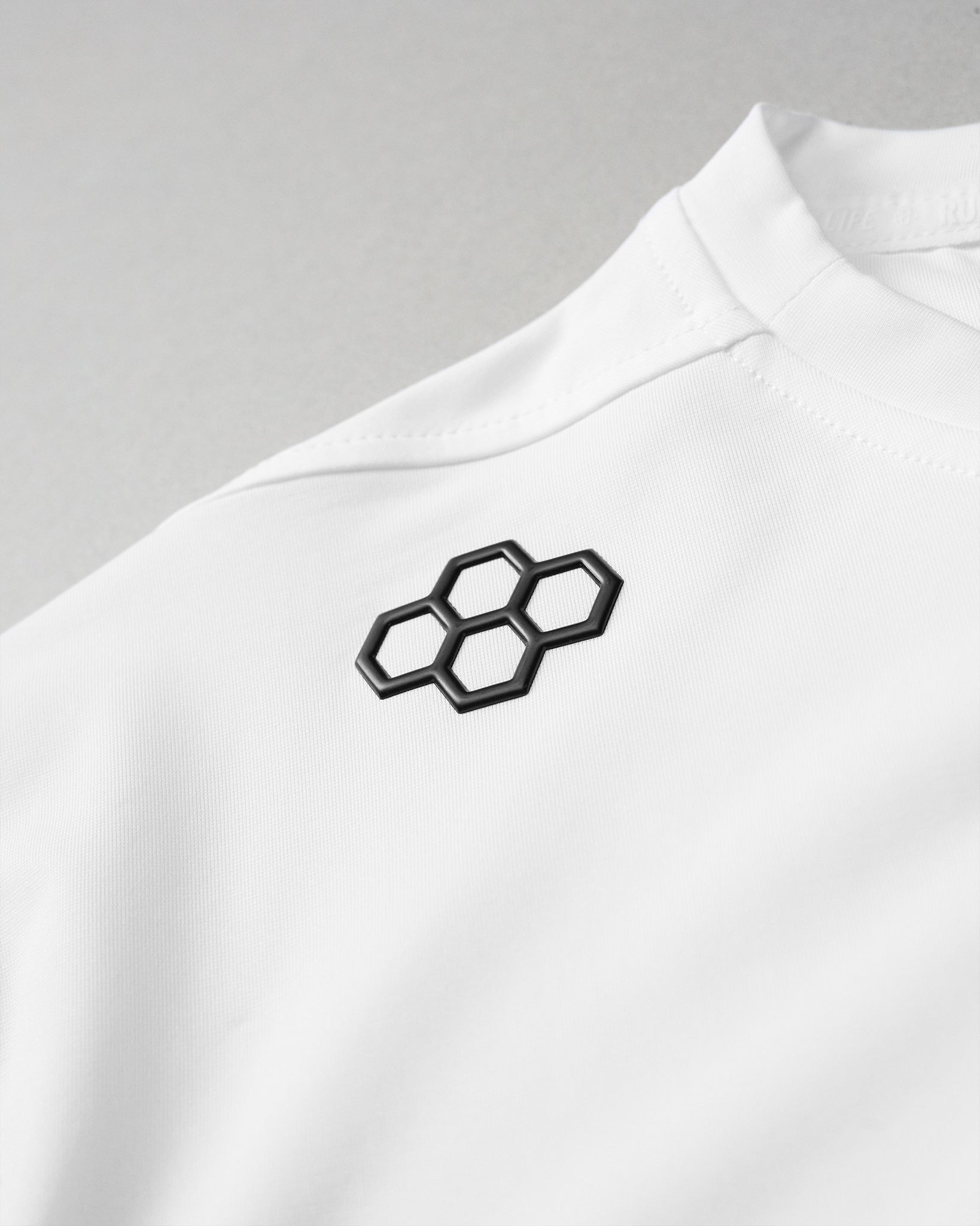 A close-up of the upper shoulder area of a white athletic shirt. The focus is on a raised black RUDIS hex logo made of six interlocking hexagons, located near the front collar. The fabric has a smooth texture with visible stitching along the shoulder seam.