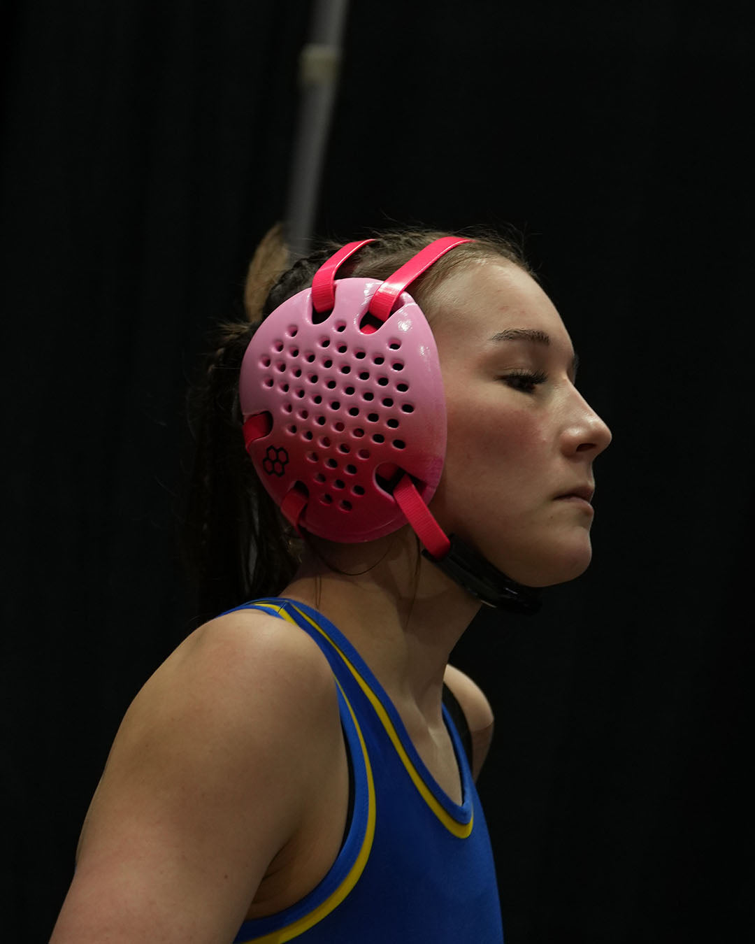 Person wearing a blue wrestling singlet and pink headgear against a black background