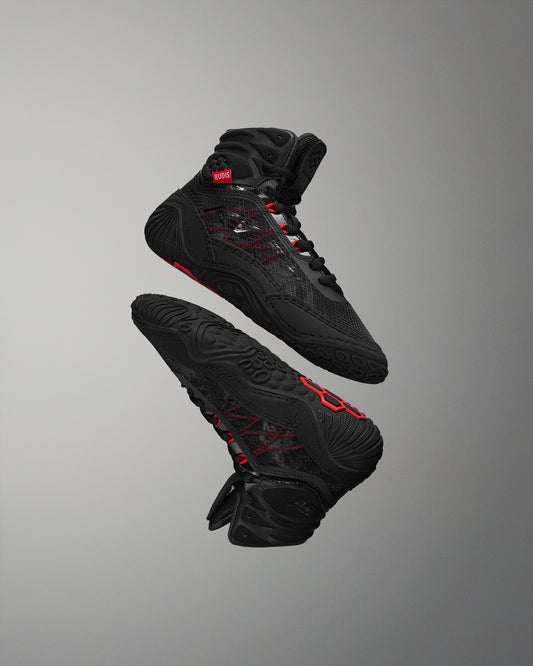 A pair of high-top black athletic shoes with red accents floats against a gray background, showcasing a sleek design and functional textured sole.