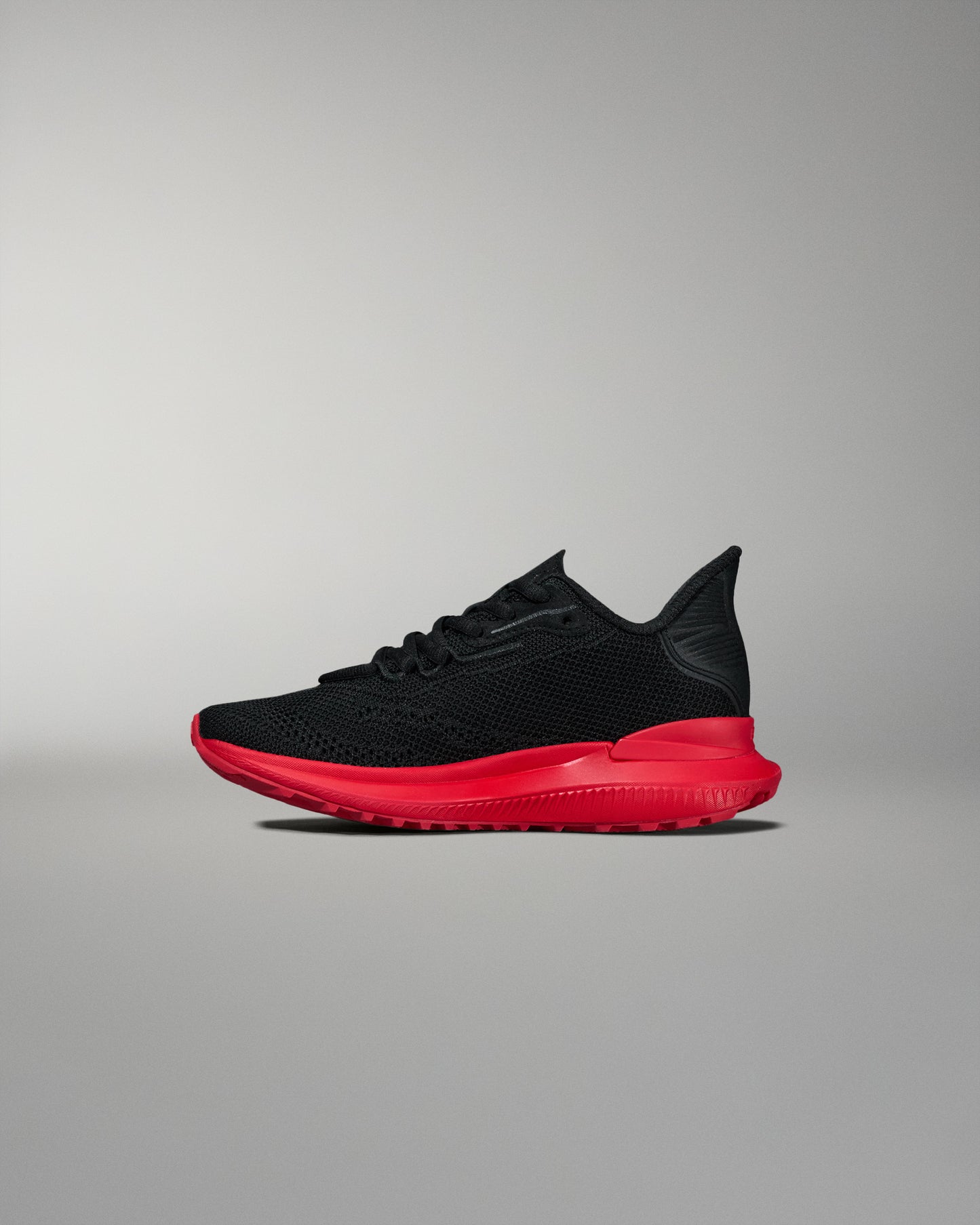 Side view of a black RUDIS training shoe with a solid red midsole and outsole, showing a clean mesh upper without visible logos. Displayed on a neutral gray background.