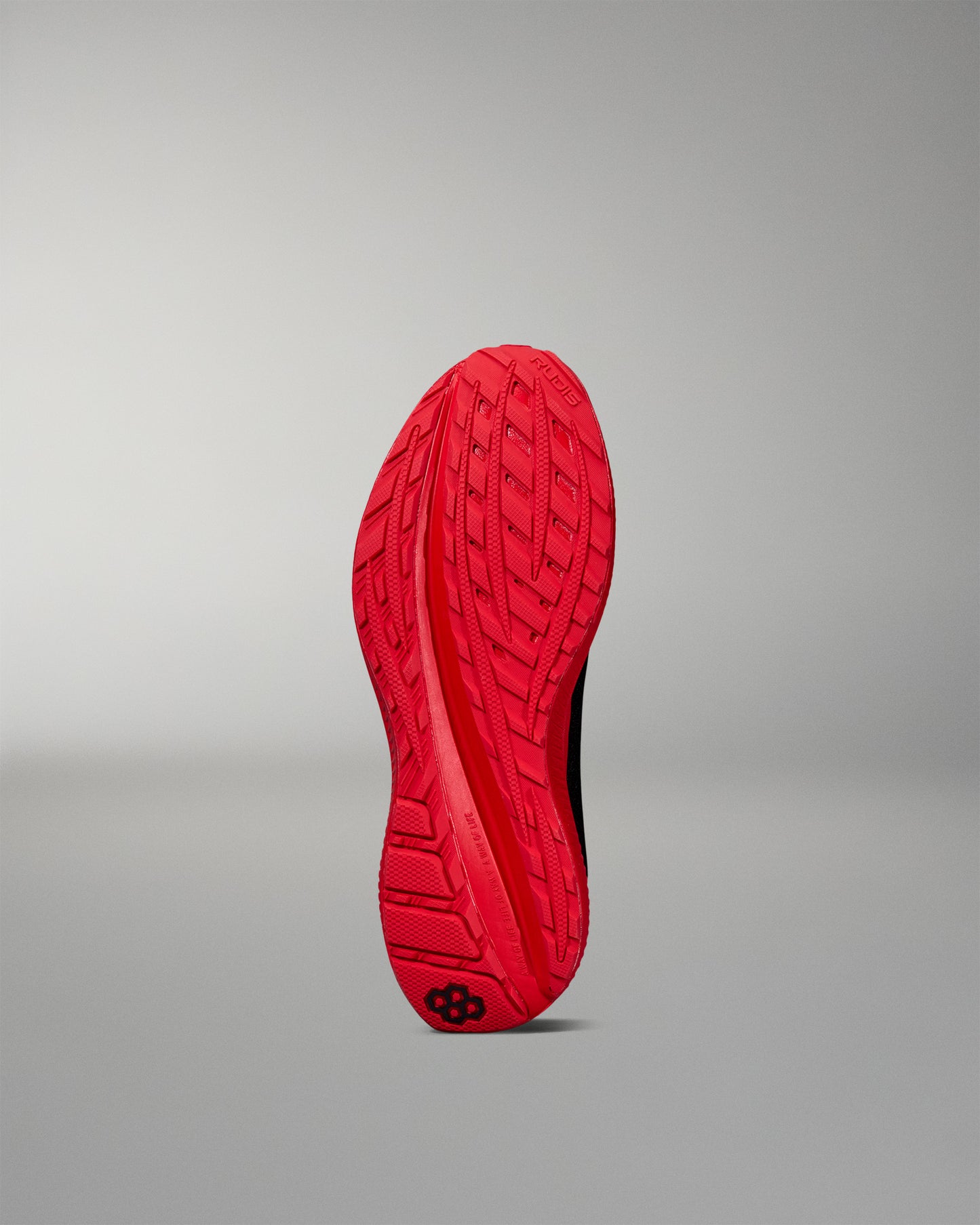 Bottom view of a red RUDIS training shoe outsole featuring a textured tread pattern and a black hexagon logo near the heel. Displayed on a neutral gray background.