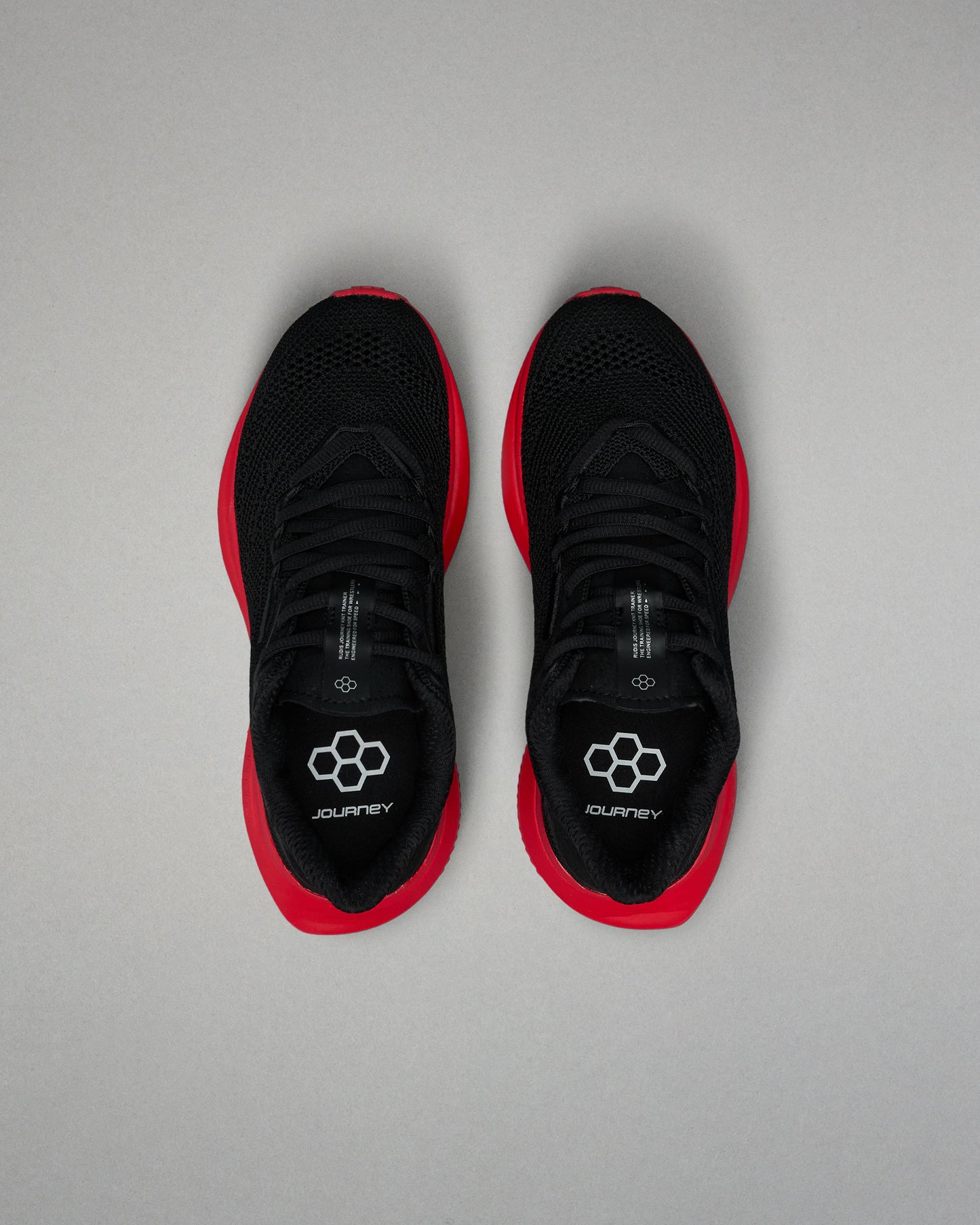 Top view of a pair of black RUDIS training shoes with red midsoles and black laces. The insoles display a white hexagon logo and the word ‘JOURNEY.’ Shoes are positioned side by side on a light gray background.