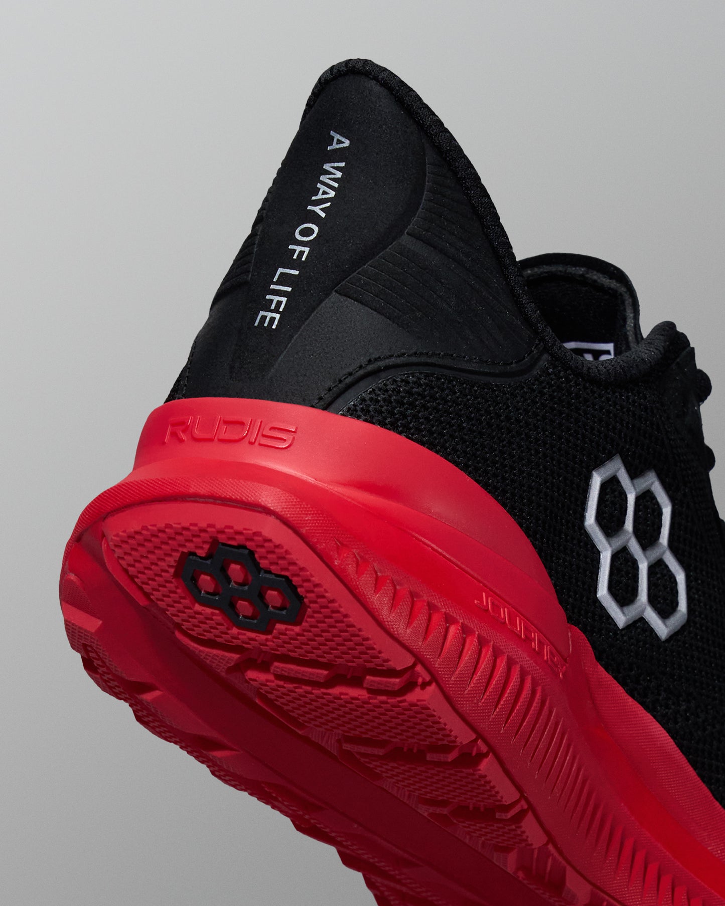 Close-up rear angle of a black RUDIS training shoe with a red outsole and midsole. Features the phrase ‘A WAY OF LIFE’ on the black heel tab, white hexagon logo on the side, and RUDIS branding on the sole.