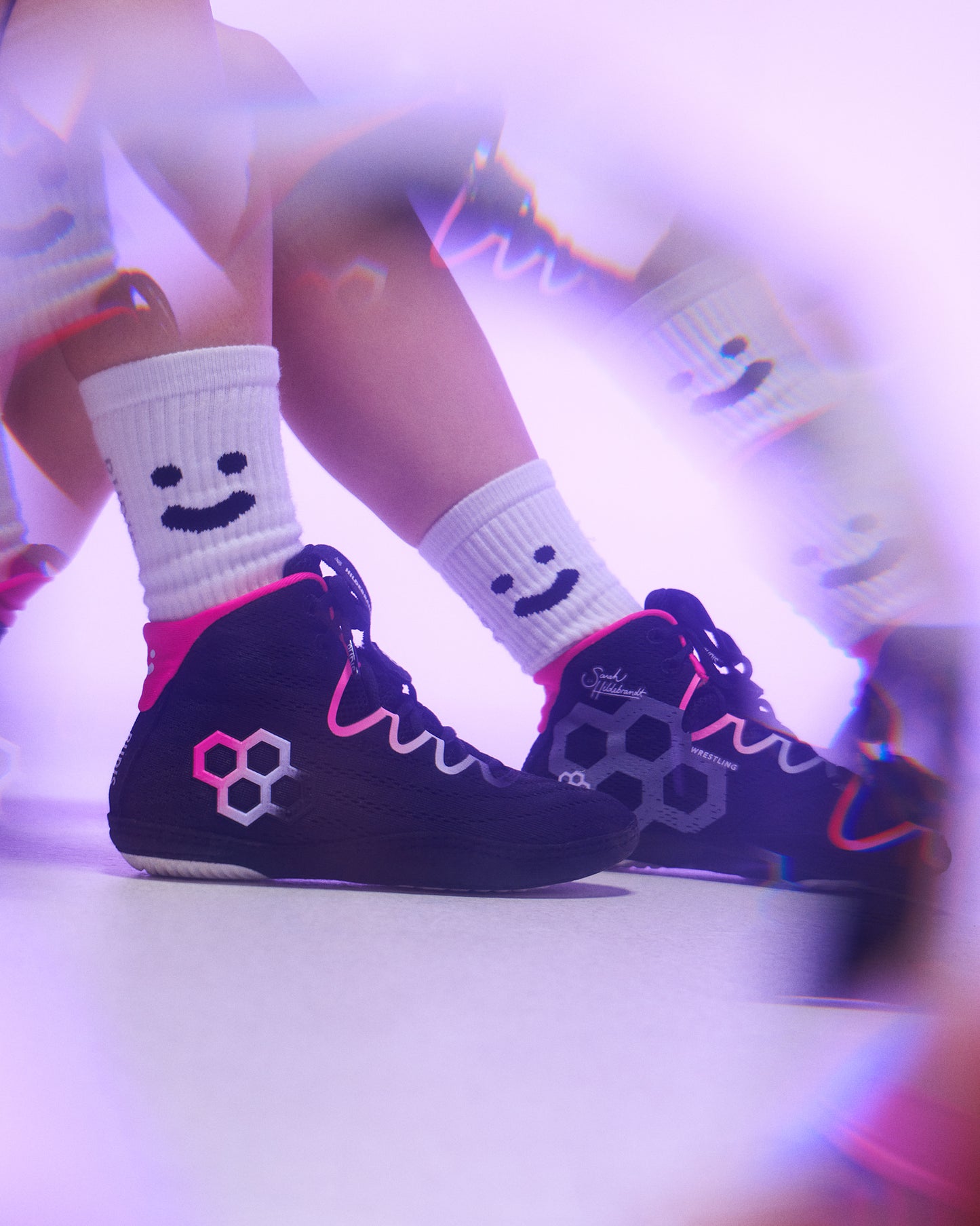 Black high-top Wrestling shoes with pink and white graphic details, worn with white crew socks featuring a smiley face, shown in a stylized purple-lit setting.
