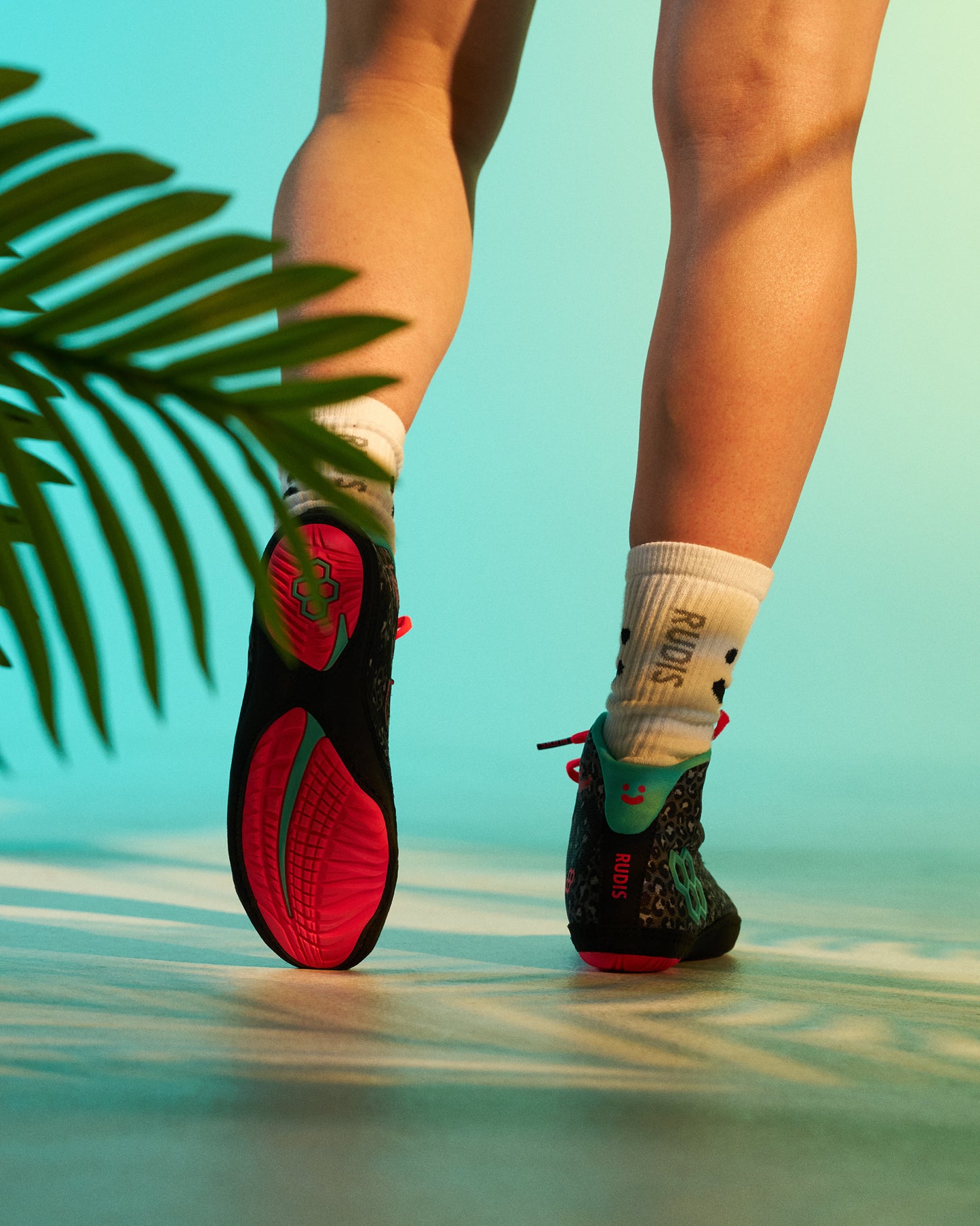Rear view of the RUDIS Hildebrandt 2.0 Girls Wrestling Shoes in Black Leopard, showcasing the bright pink outsole, teal heel accents, leopard-print upper, and RUDIS crew socks in a tropical-inspired studio setting