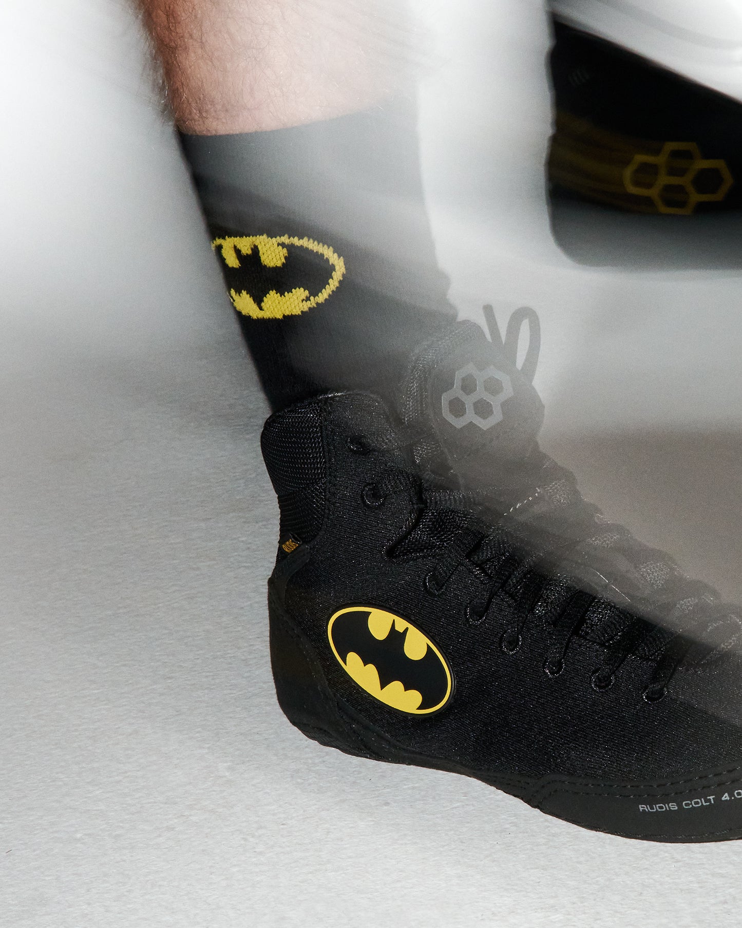 A close-up of a black high-top wrestling shoe with a yellow Batman logo on the side, worn with a black sock featuring a yellow Batman logo. The shoe has a textured knit upper and black laces, shown against a light gray background with a motion blur effect.