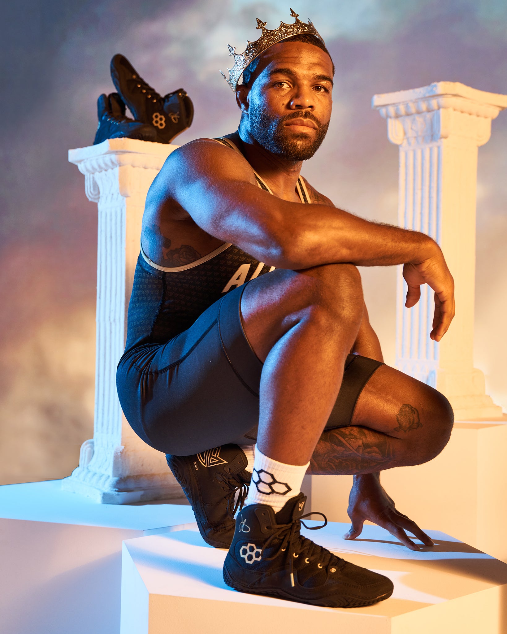 A muscular athlete in a black outfit and a crown poses confidently showcasing wrestling shoes and a striking background