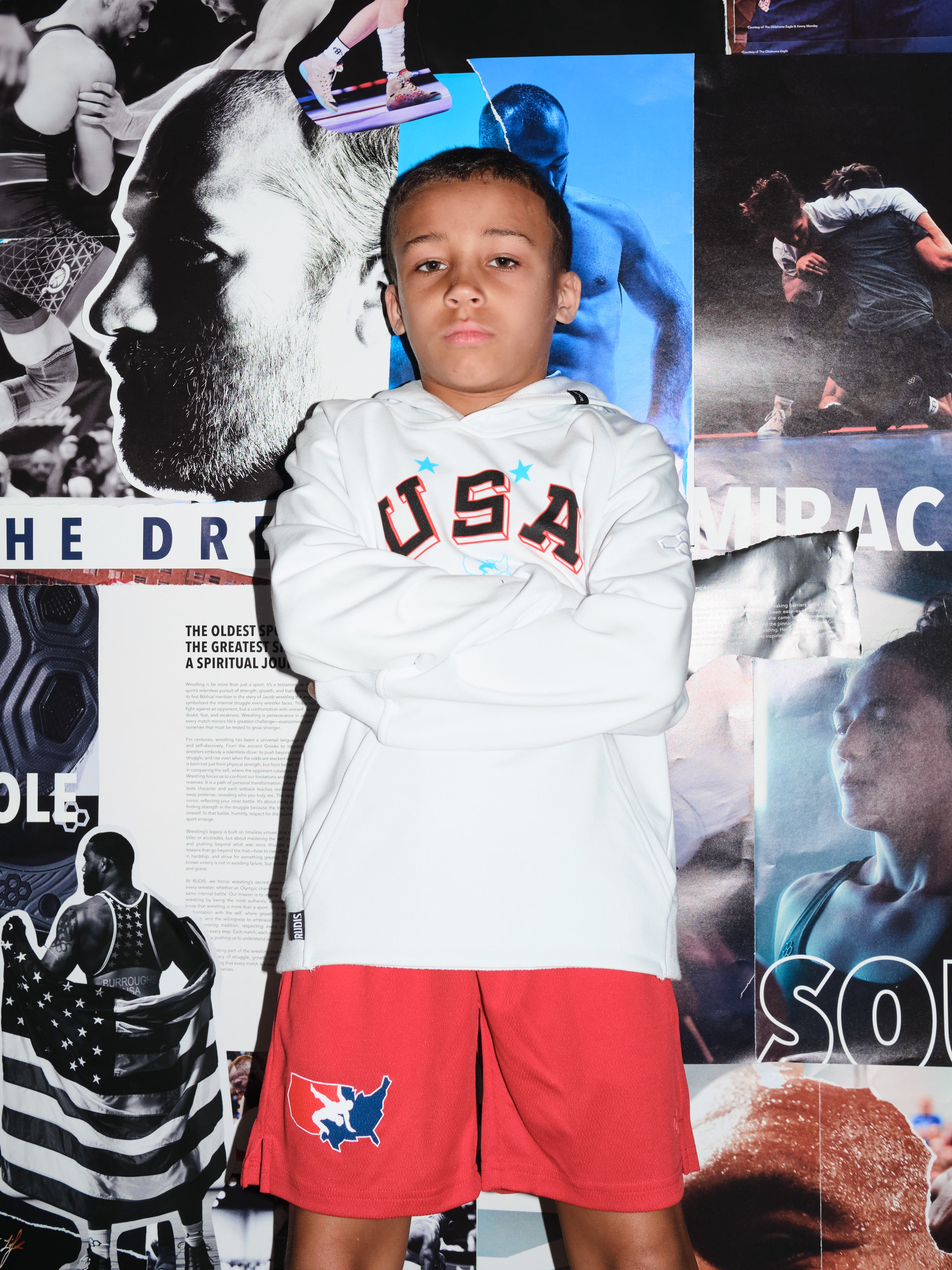 Beacon Burroughs, youth wrestler and son of Olympic champion Jordan Burroughs, wearing a white USA Wrestling hoodie and red USA Wrestling shorts by RUDIS, standing confidently in front of a wrestling mural — representing the next generation of wrestling.