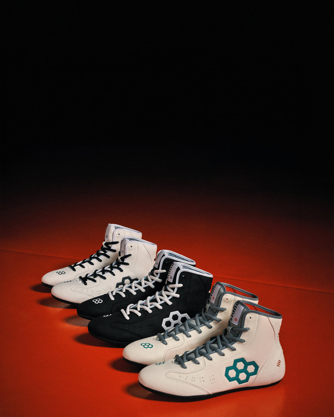 Image of 3 pairs of RUDIS International wrestling shoes sitting side by side on an orange and black wrestling mat. 