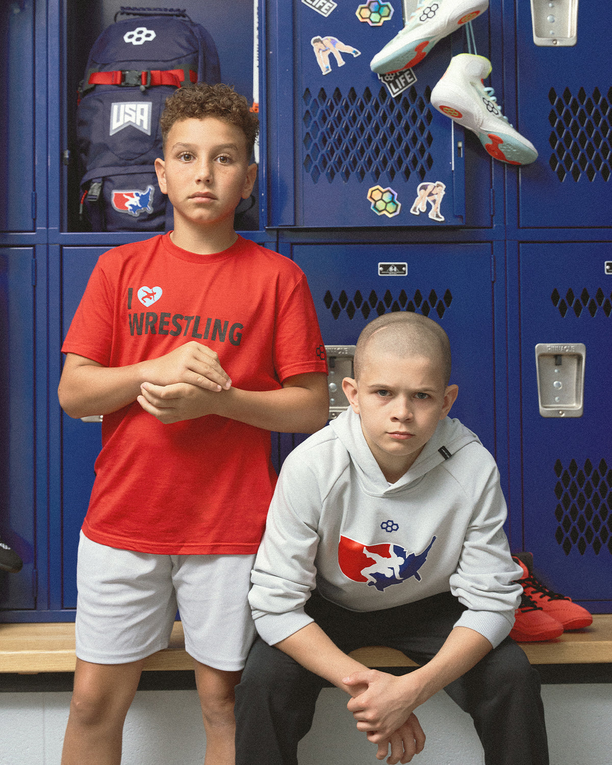 Two youth wrestlers in a locker room wearing RUDIS and USA Wrestling apparel, with RUDIS wrestling shoes and gear packs in the background — showcasing wrestling and training essentials.