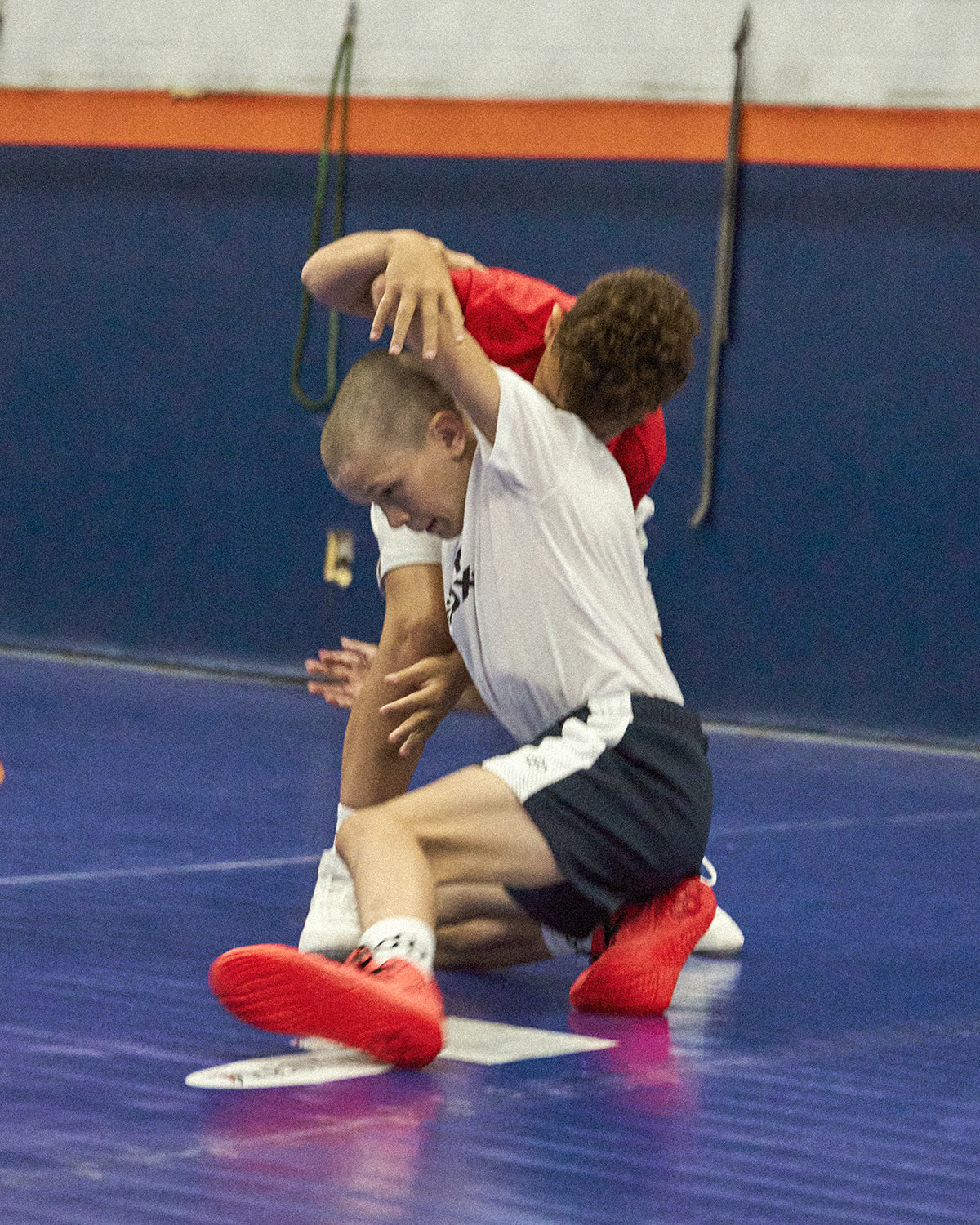 Youth wrestlers in action during practice on a blue mat, wearing RUDIS gear and red wrestling shoes while drilling takedowns — showcasing youth wrestling dedication and training. 