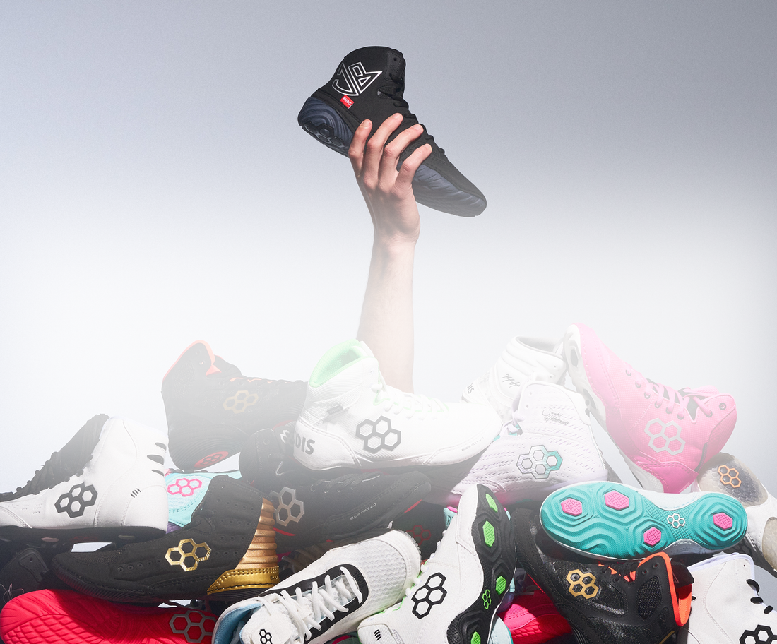 Arm holding RUDIS JB1 wrestling shoes in the black metal colorway. The image also features a stack of other RUDIS performance footwear including JB1, JB Ultra, JB-UltraLITE, Colt 4.0, International, Alpha 2.0, and Hildebrandt models.