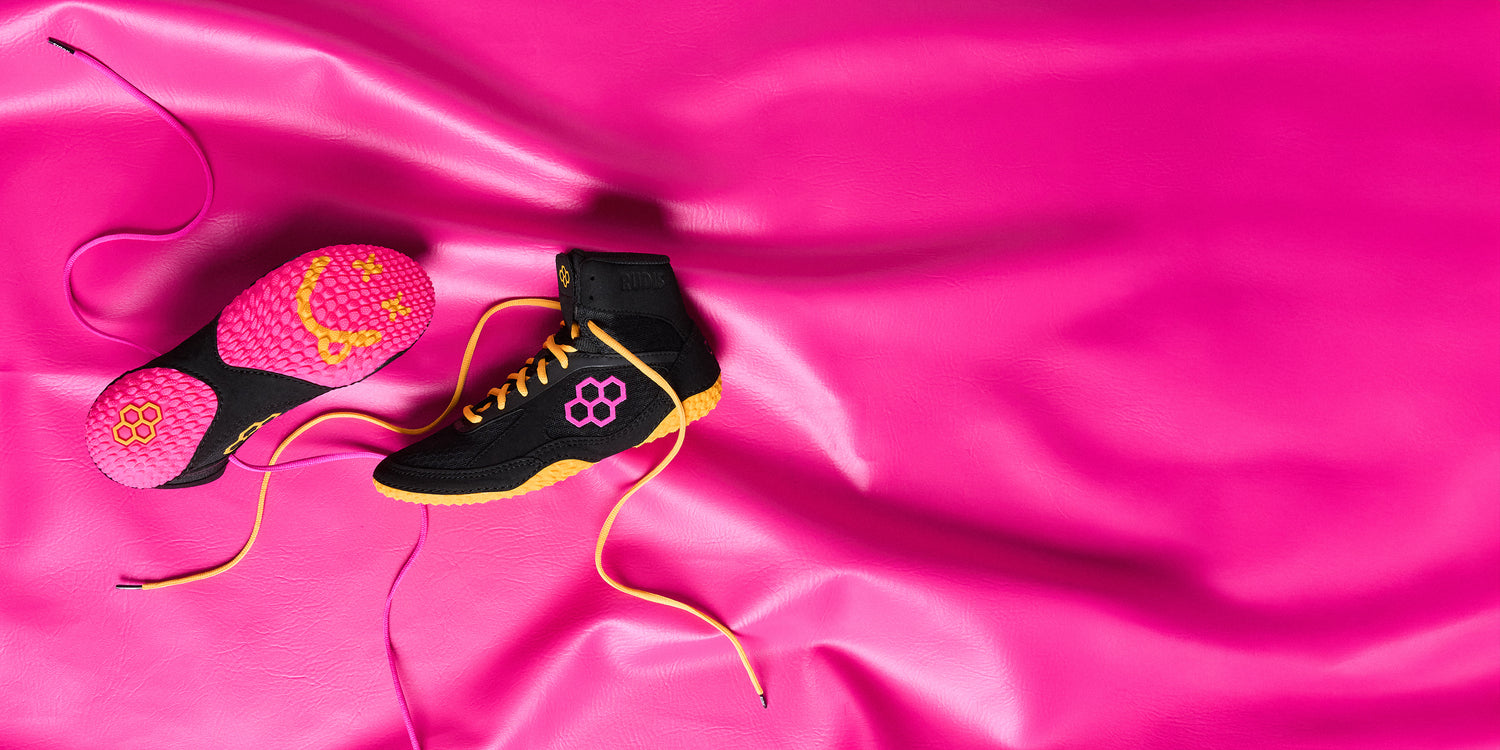 RUDIS Alpha 2.0 wrestling shoes in Happy 2.0 colorway with black, pink, and gold accents, highlighting bold design, grip traction, and lightweight performance