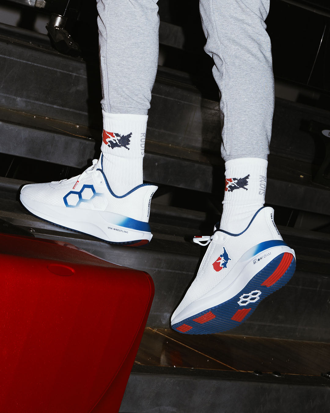 White RUDIS sneakers with blue and red accents worn by a person sitting on a car.