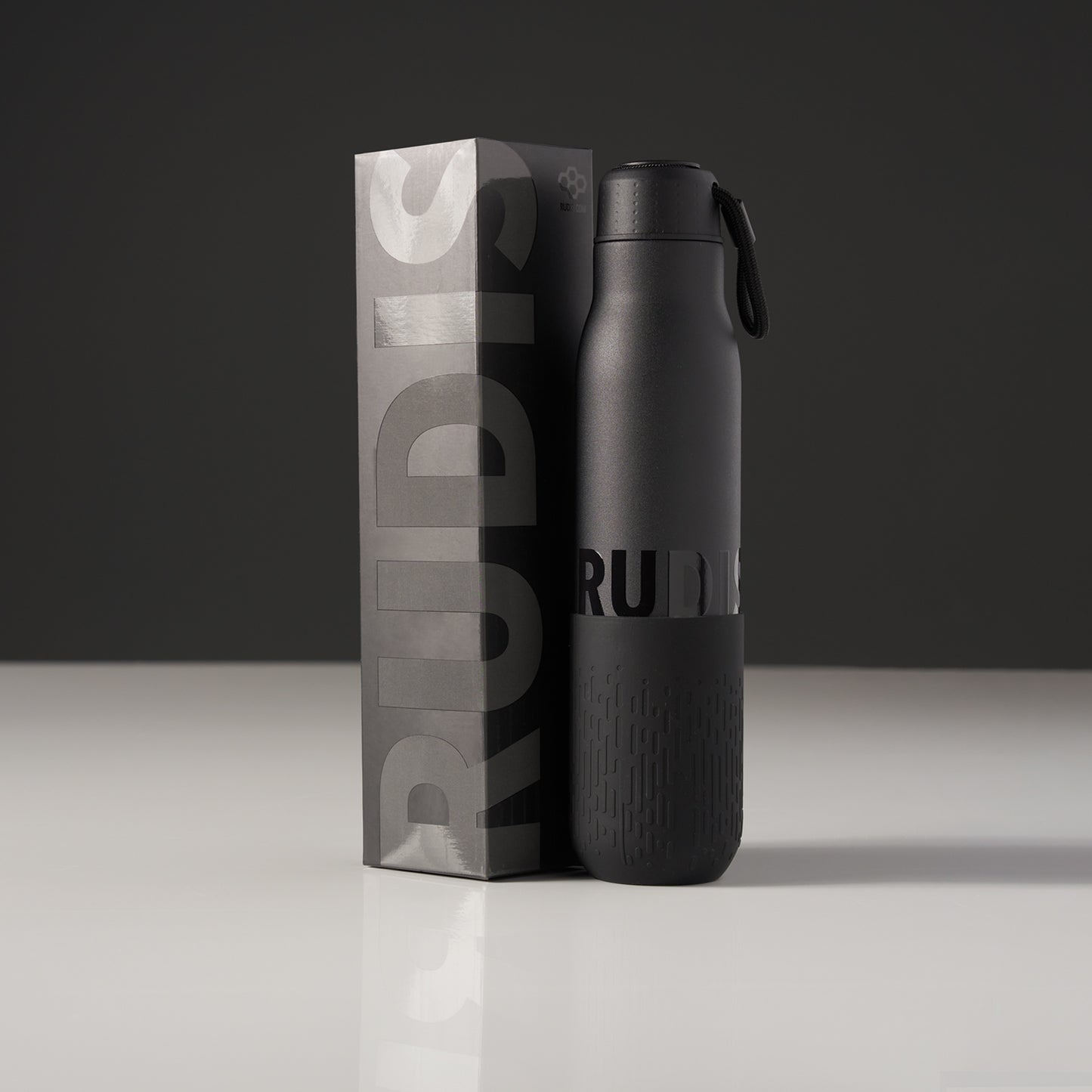 A sleek black water bottle with a textured grip standing next to its packaging featuring bold typography that reads RUDIS