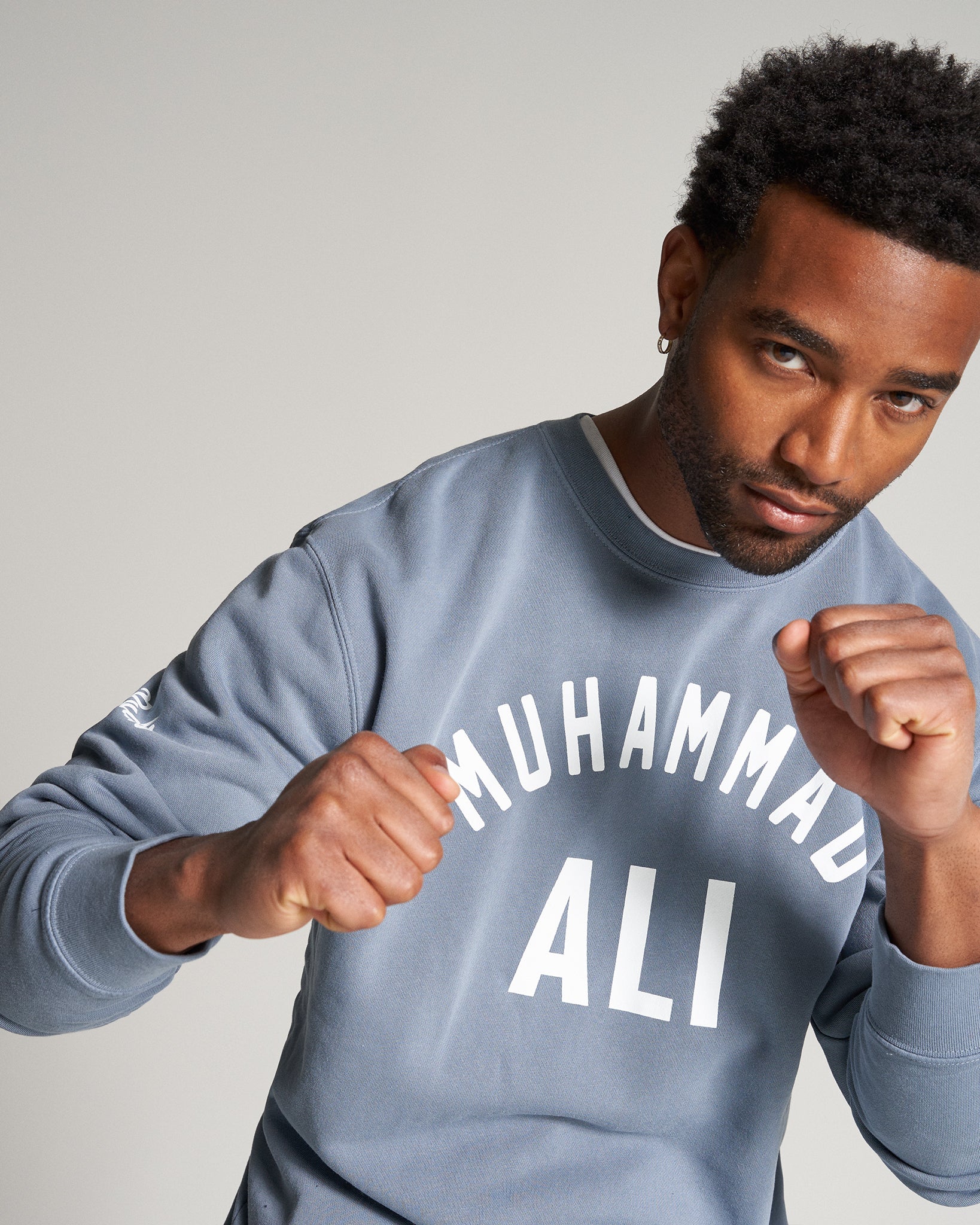 A man wearing a gray sweatshirt featuring the words MUHAMMAD ALI in bold white letters showcasing a confident stance with clenched fists against a neutral background