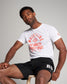 A male model poses confidently in a casual outfit consisting of a white t-shirt with a bold red wrestling graphic and black athletic shorts seated on a wooden stool against a light grey background