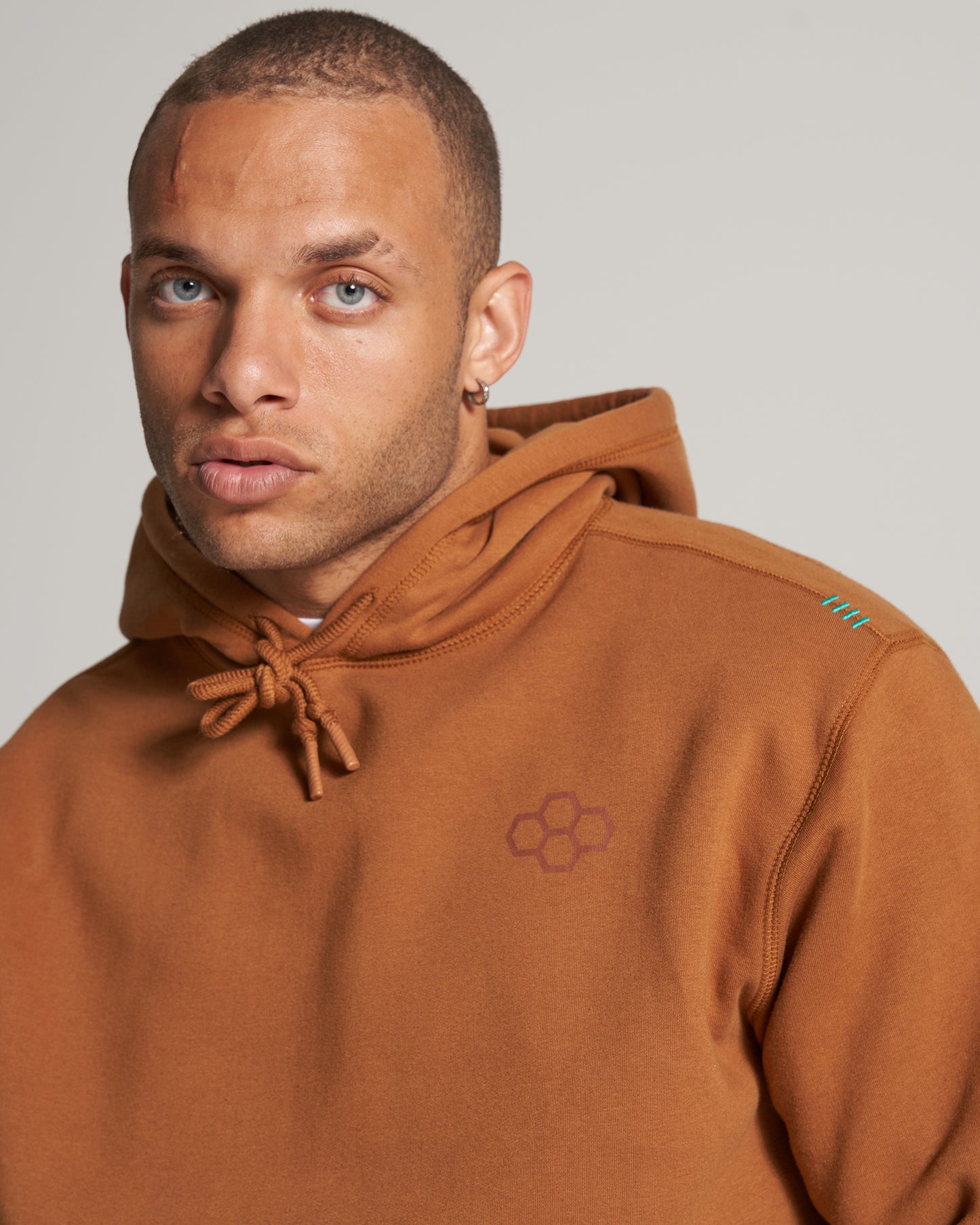A close-up portrait of a man wearing a brown hoodie featuring a subtle logo and bright blue accents with a serious expression and skin imperfections visible