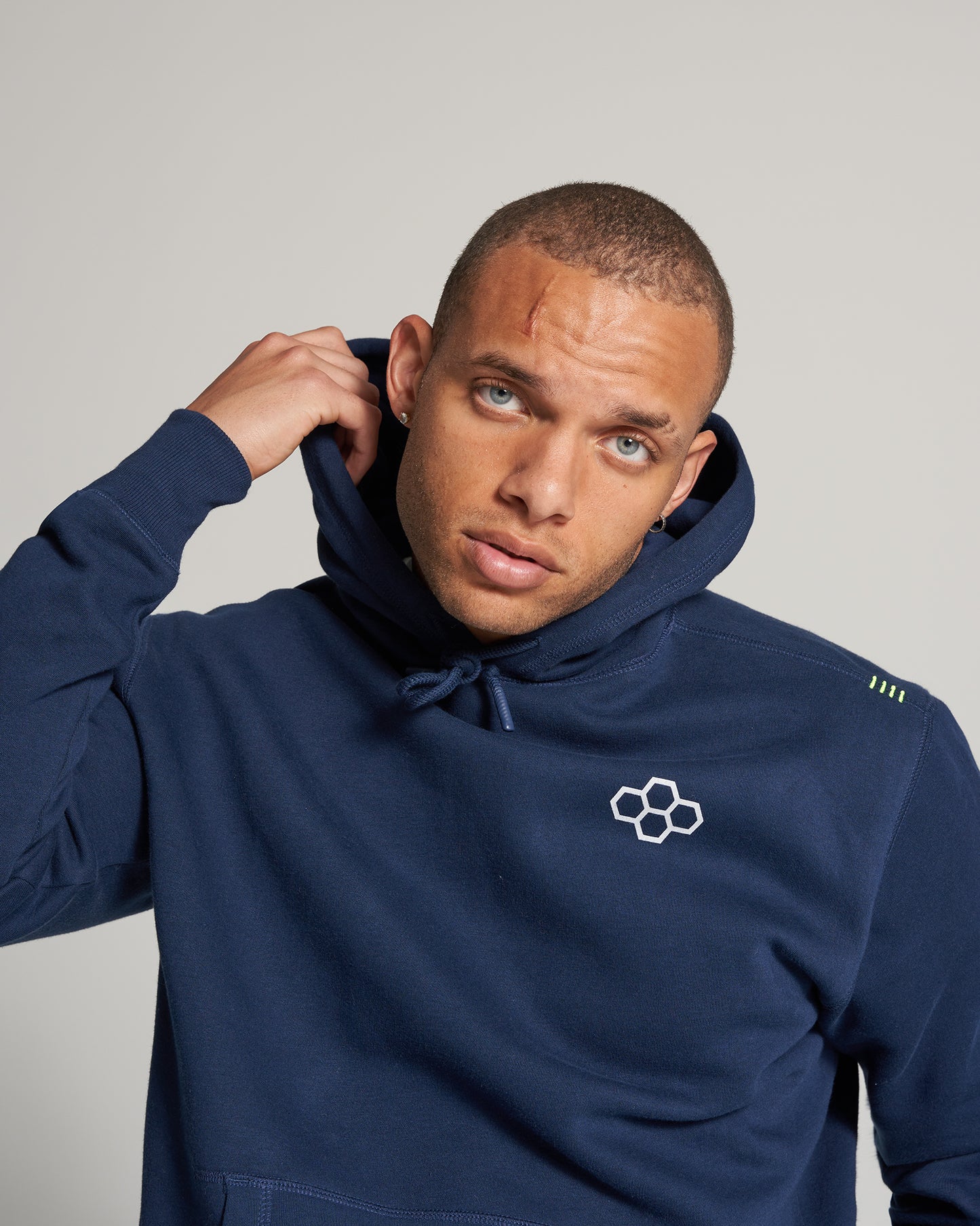 A man wearing a navy blue hoodie with a logo on the chest showcasing a casual style complemented by a serious expression and a visible scar on his forehead