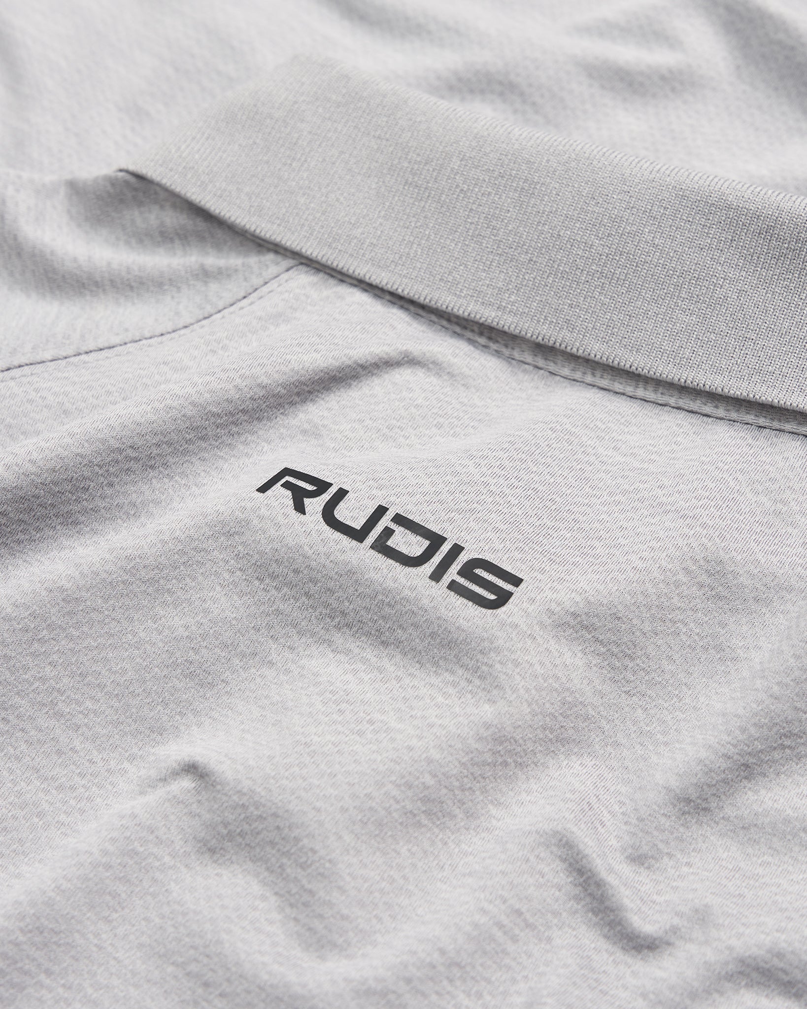 A close-up view of a light gray polo shirt featuring a black RUDIS logo on the collar area emphasizing a stylish and sporty aesthetic