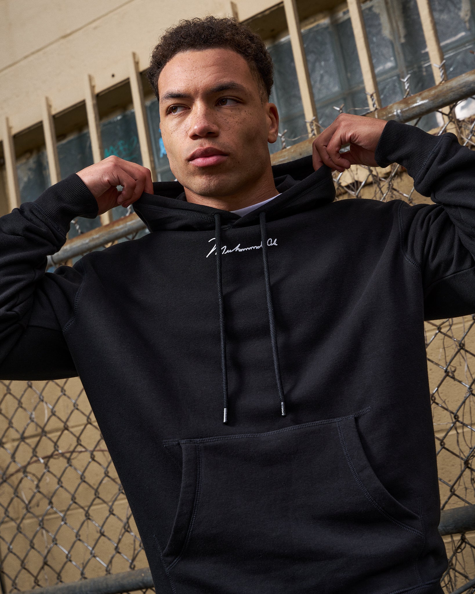 Person wearing a black hoodie with text, standing against a chain-link fence.