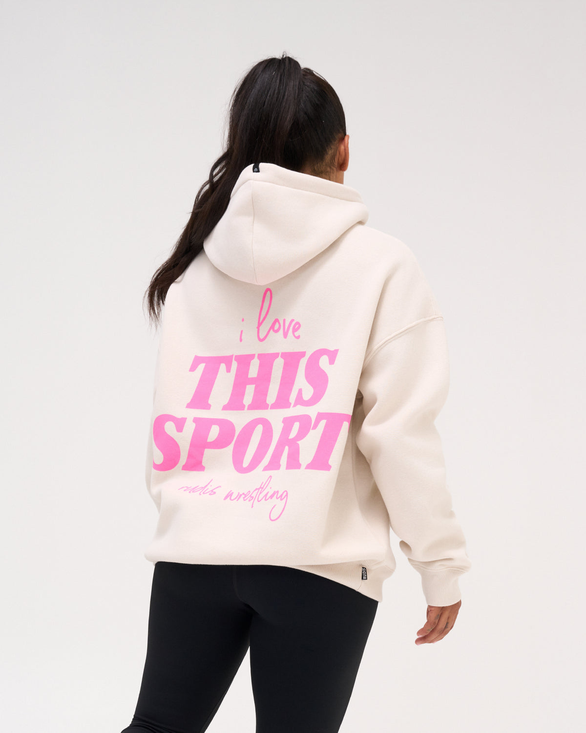 Back view of a female model wearing the RUDIS Love This Sport Relaxed Comfort Fleece Hoodie in Bone, featuring a bold pink I Love This Sport wrestling graphic.