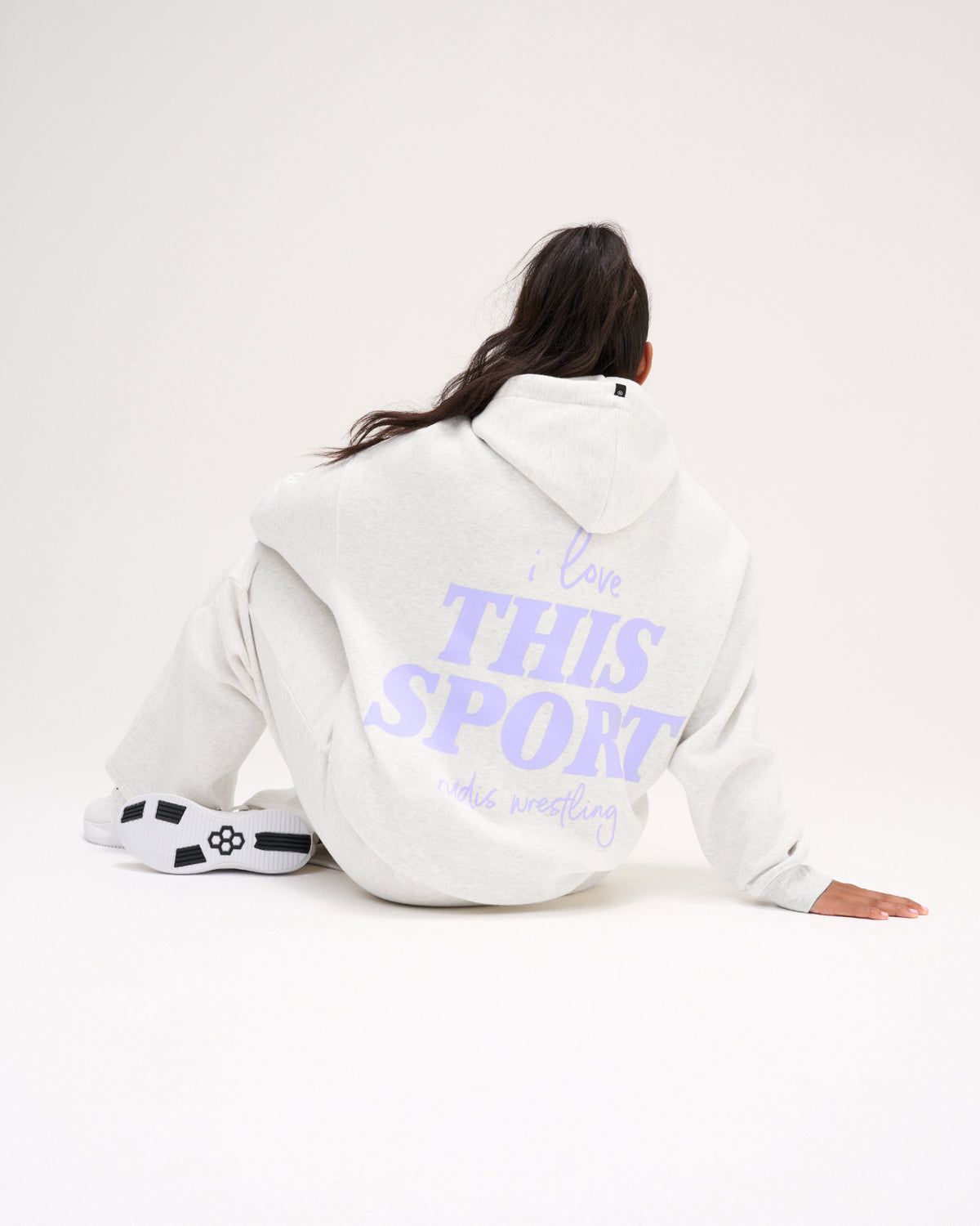 Seated female model showing the back of the RUDIS Love This Sport Heather Silver fleece hoodie with a large lavender I Love This Sport wrestling graphic.